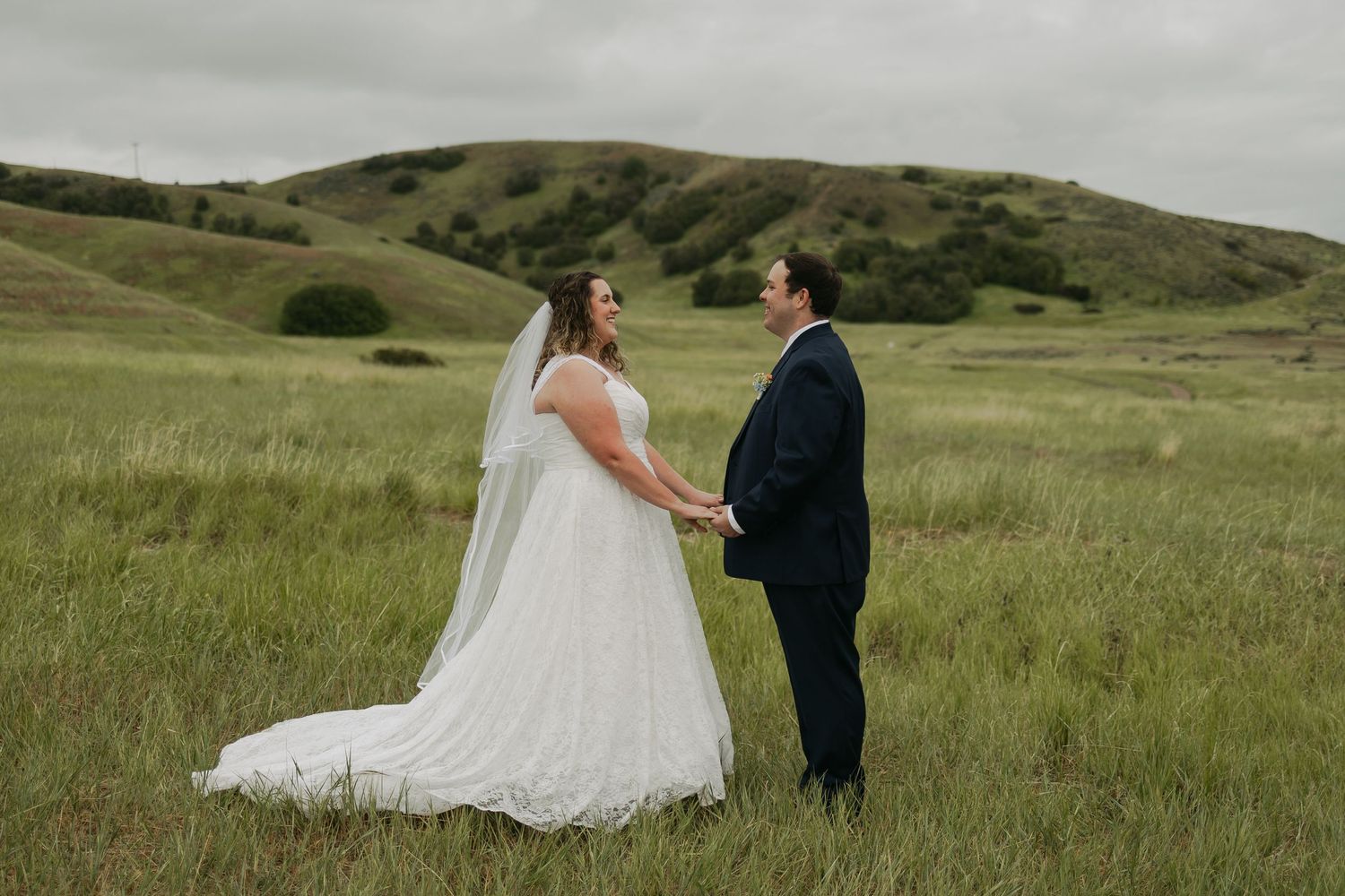 Bride and groom holding hands in a lush green meadow with rolling hills, sharing a romantic moment on their wedding day.