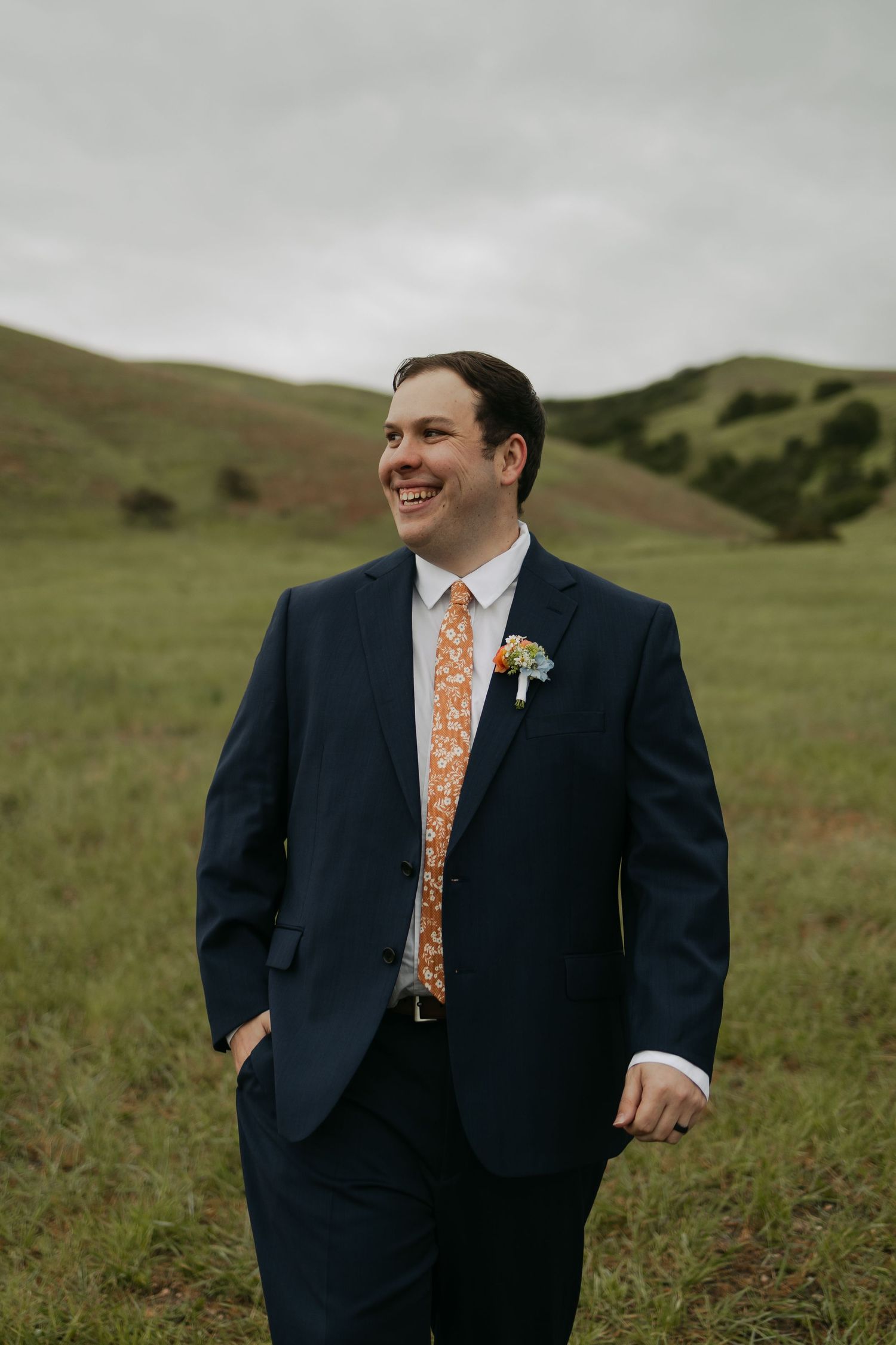 Smiling groom in navy suit with orange floral tie and boutonniere standing in green meadow with rolling hills.