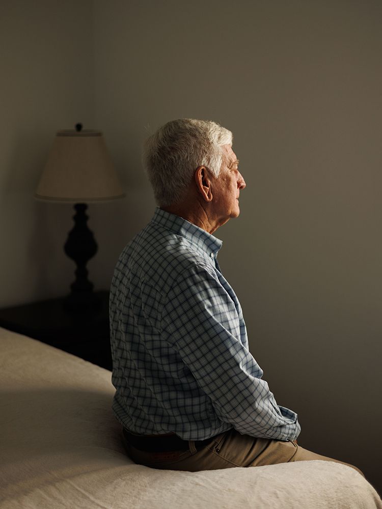 Elderly man sitting alone on bed edge in dimly lit bedroom, viewed from behind, appearing contemplative.