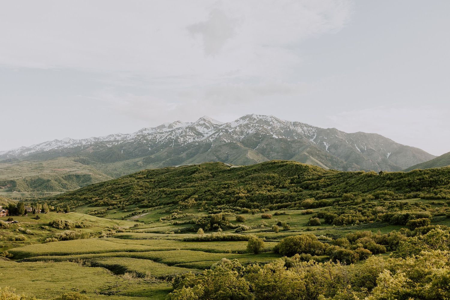 Snow-capped mountain surrounded by lush green hills and farmland under a cloudy sky.