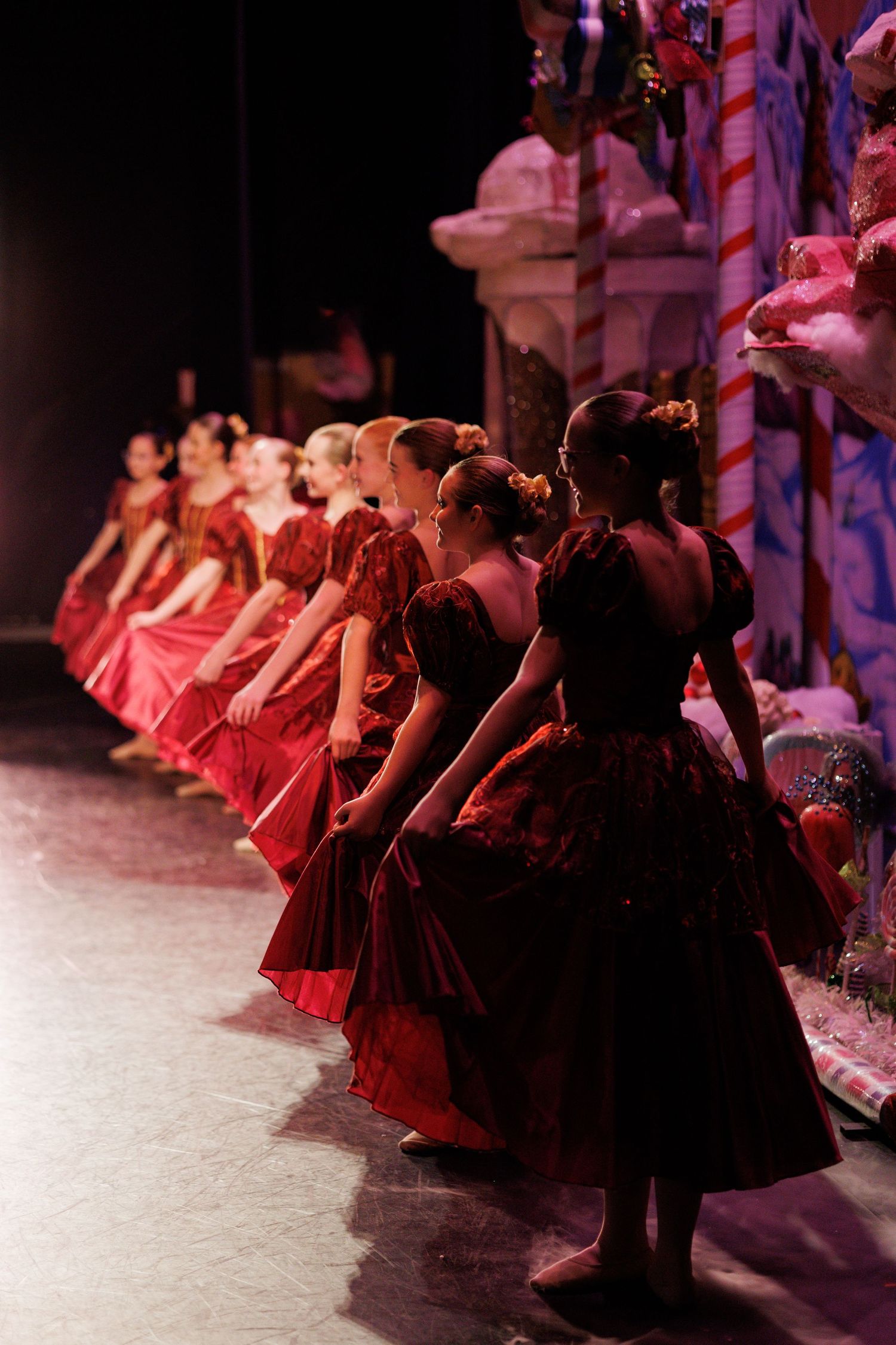 Ballet dancers in red dresses line up on stage during a performance in dramatic lighting.