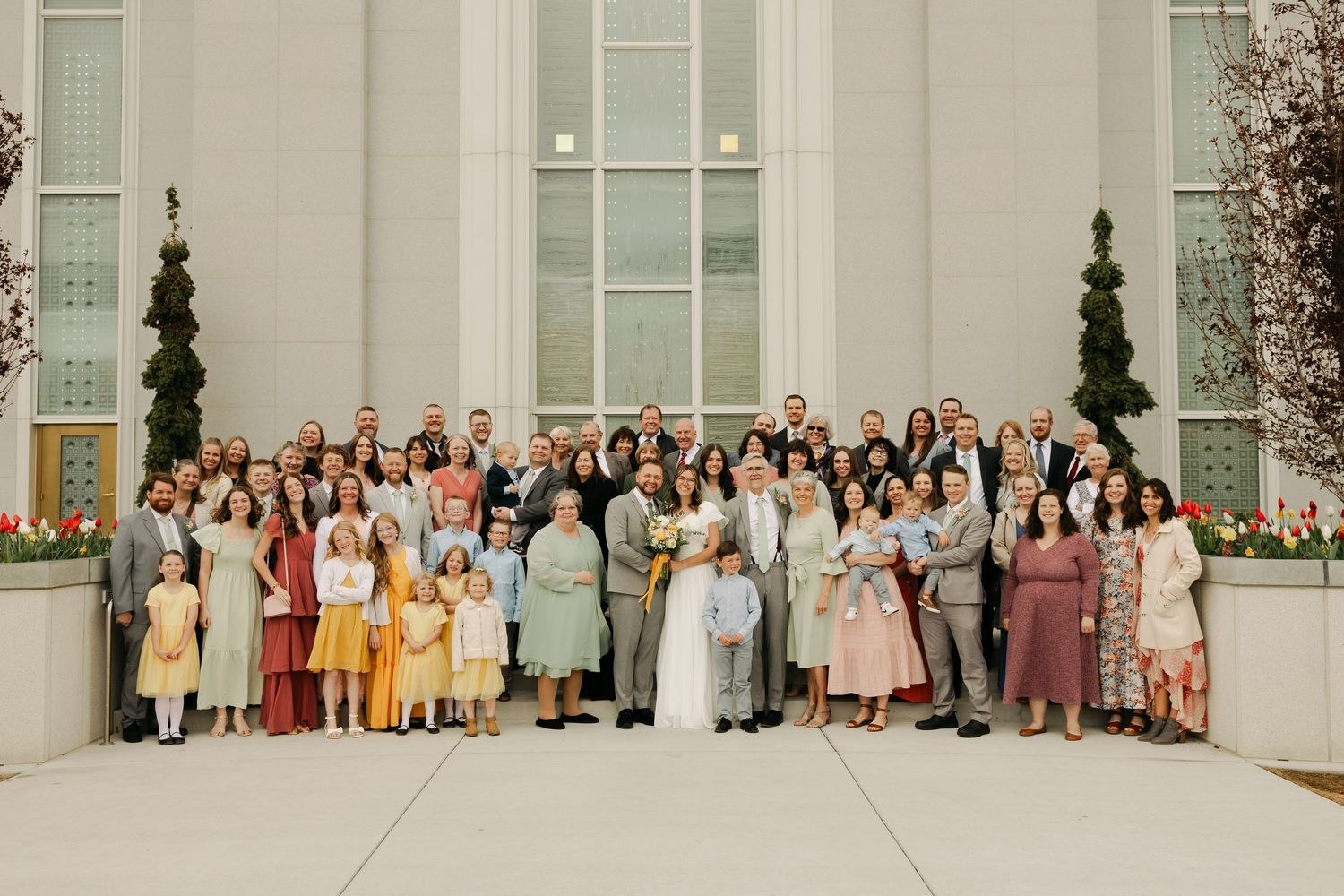 A large wedding group photo with guests in pastel attire lined up against a white building exterior.