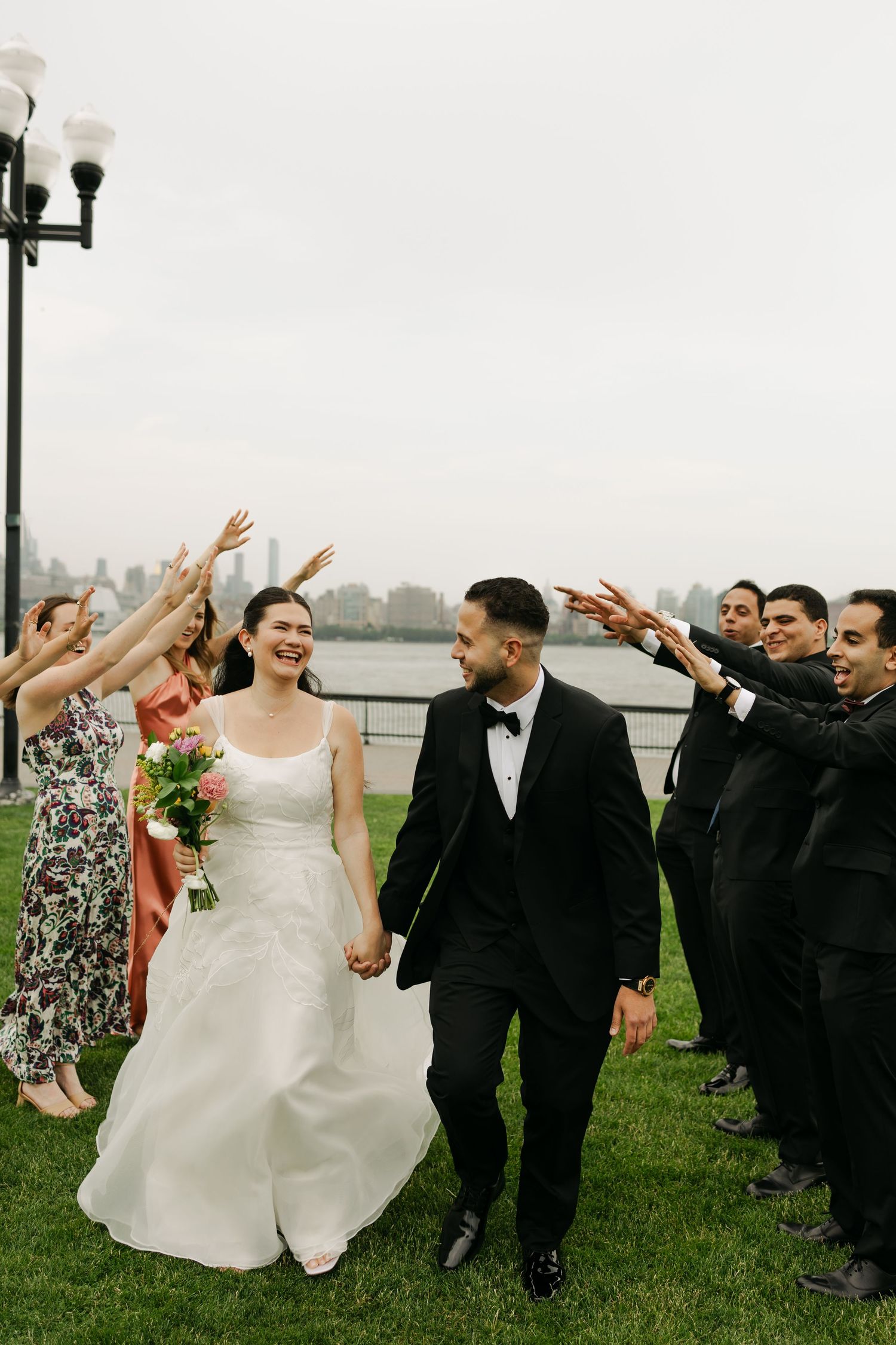 Wedding party cheers and raises arms as newlyweds walk past on waterfront path.