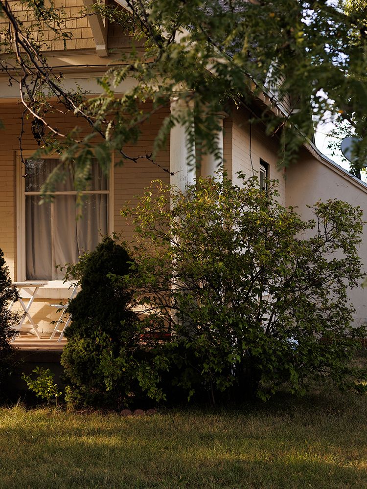 Charming yellow cottage exterior with white trim, large window, and lush green shrubs in warm sunlight.