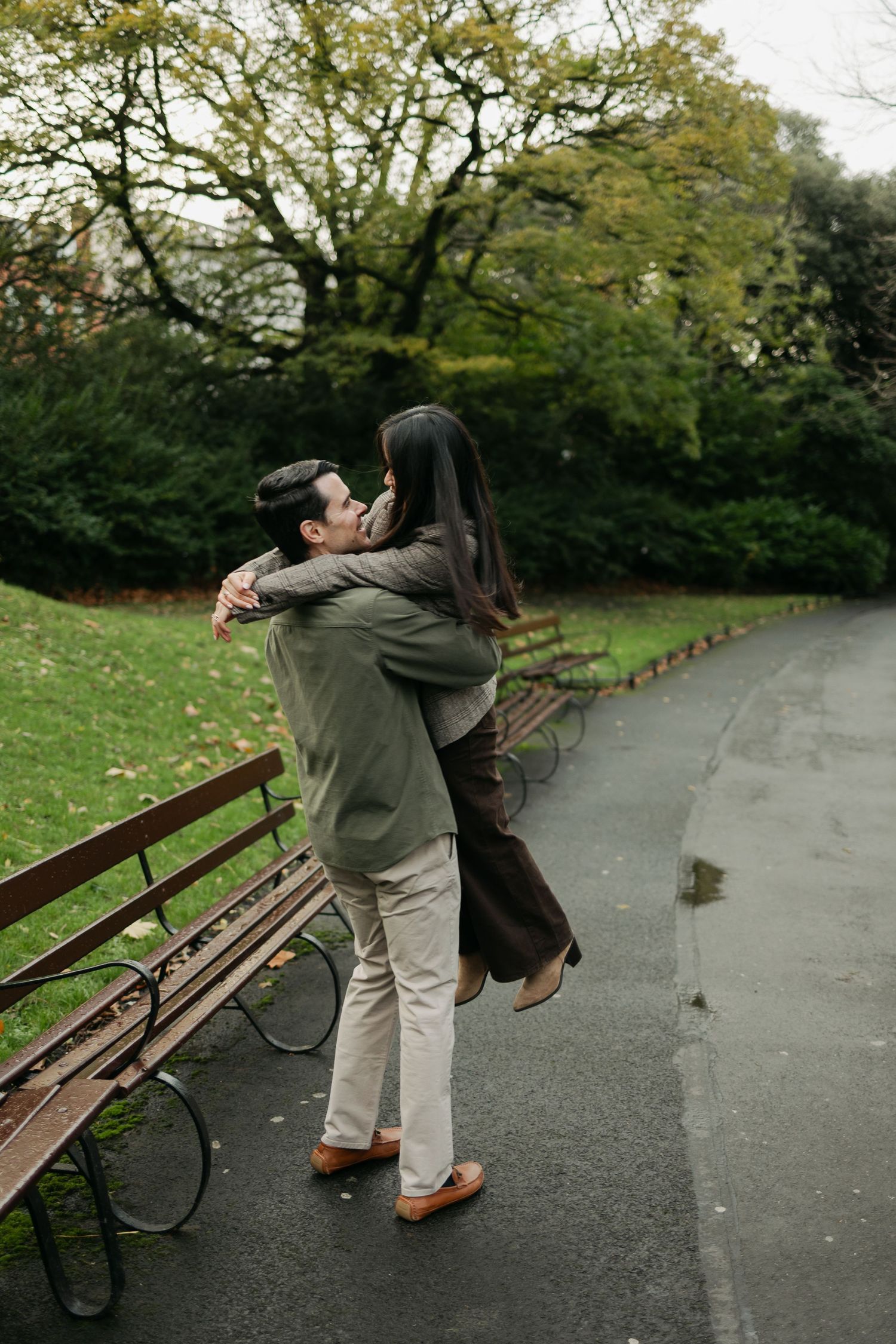 Couple embracing on a park path in autumn, man lifting woman near wooden benches with lush green trees.