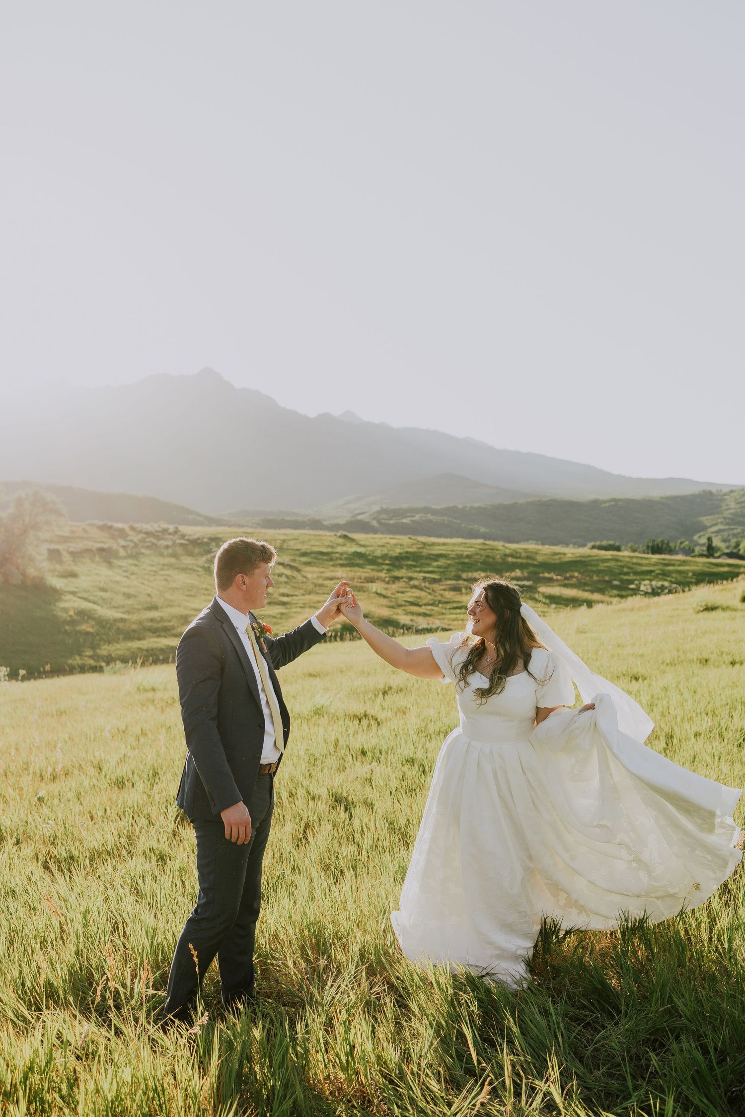 A couple dances together in a scenic mountain meadow at sunset while the bride's white dress and veil flow in the wind.