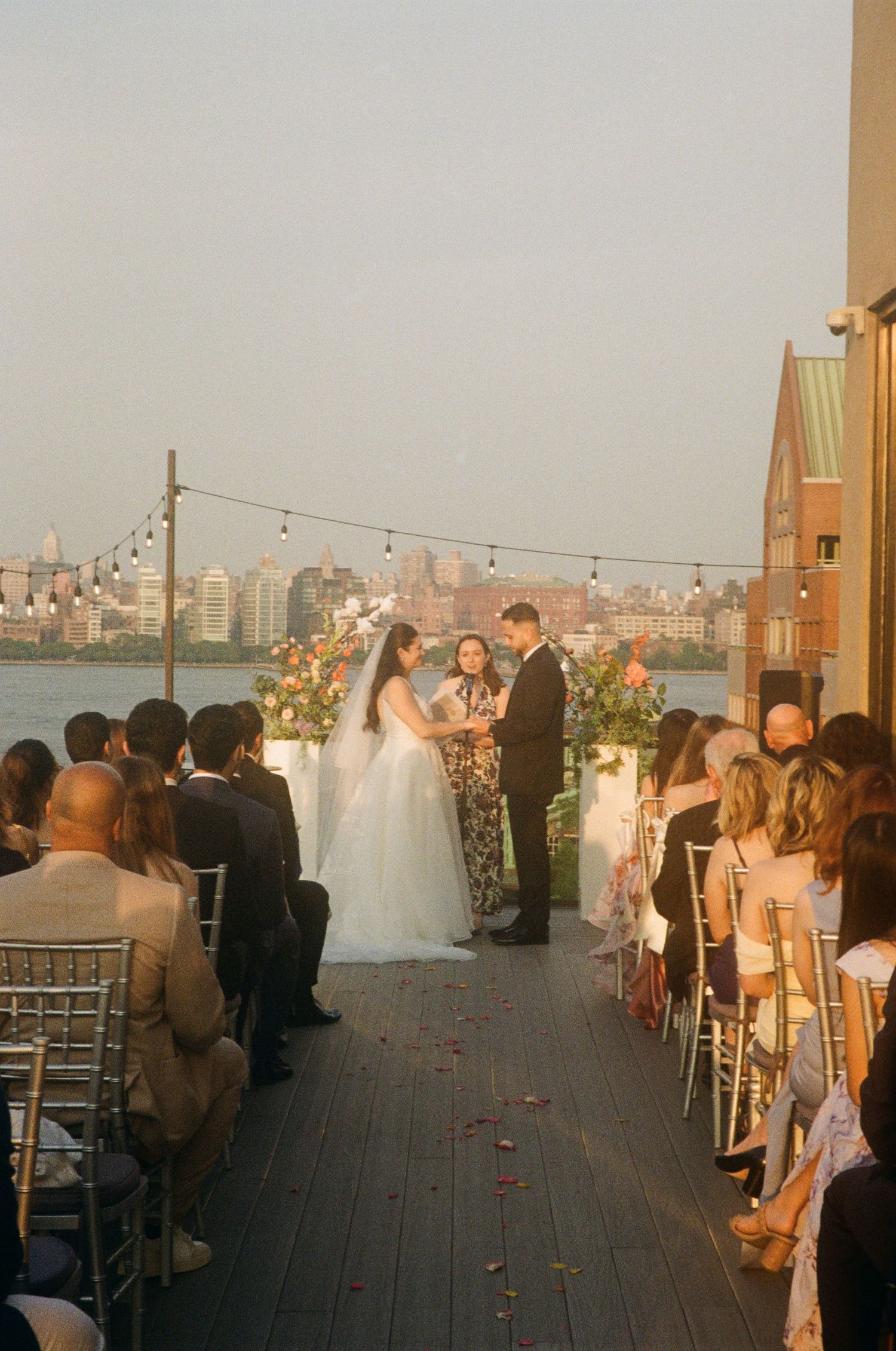 Outdoor rooftop wedding ceremony at sunset overlooking a city skyline with guests seated on both sides of the aisle.