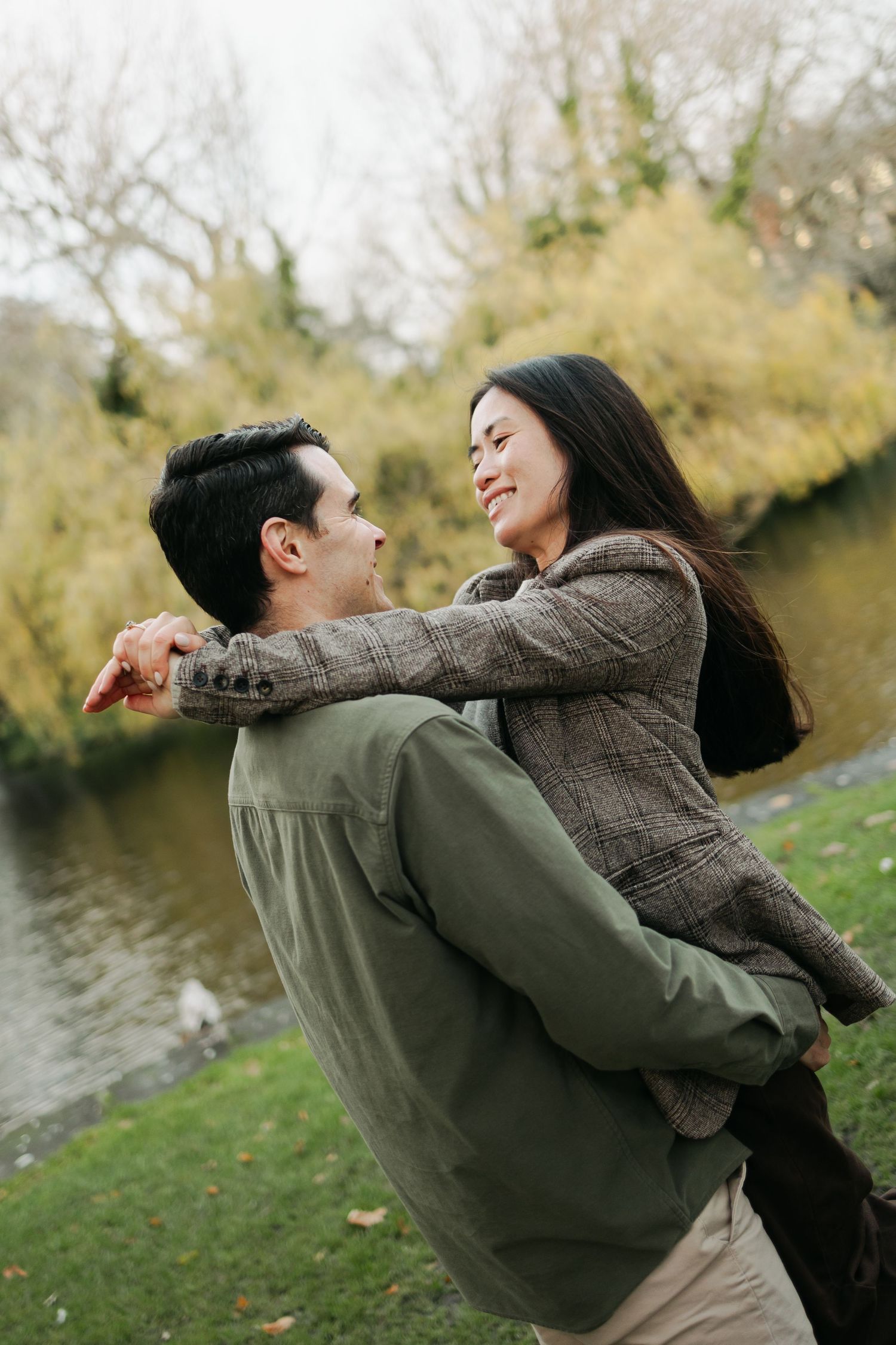 Joyful man lifting smiling woman in plaid blazer by a pond in an autumn park with willow trees.