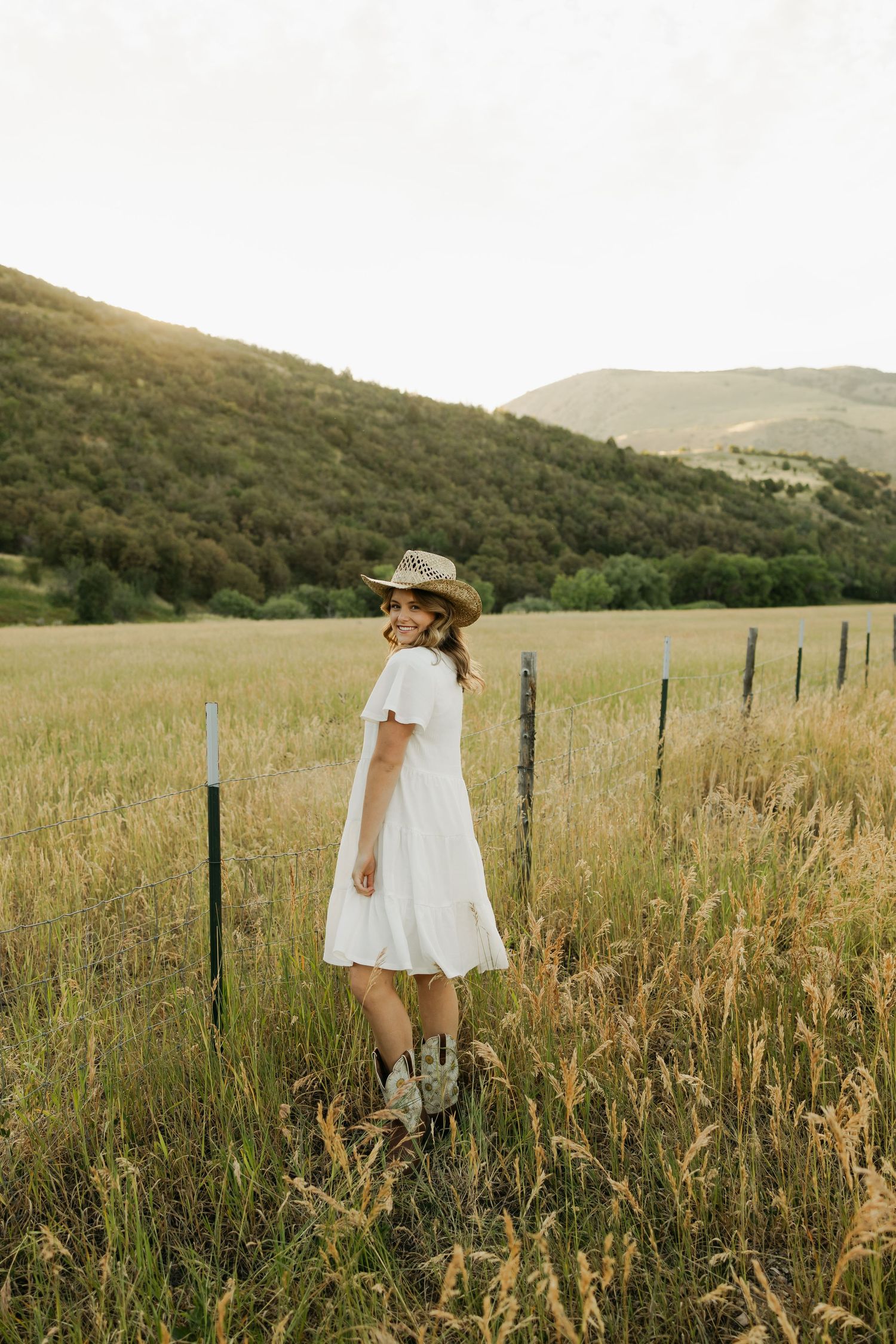 Figure in white dress standing among tall grasses with mountains and fence in background.