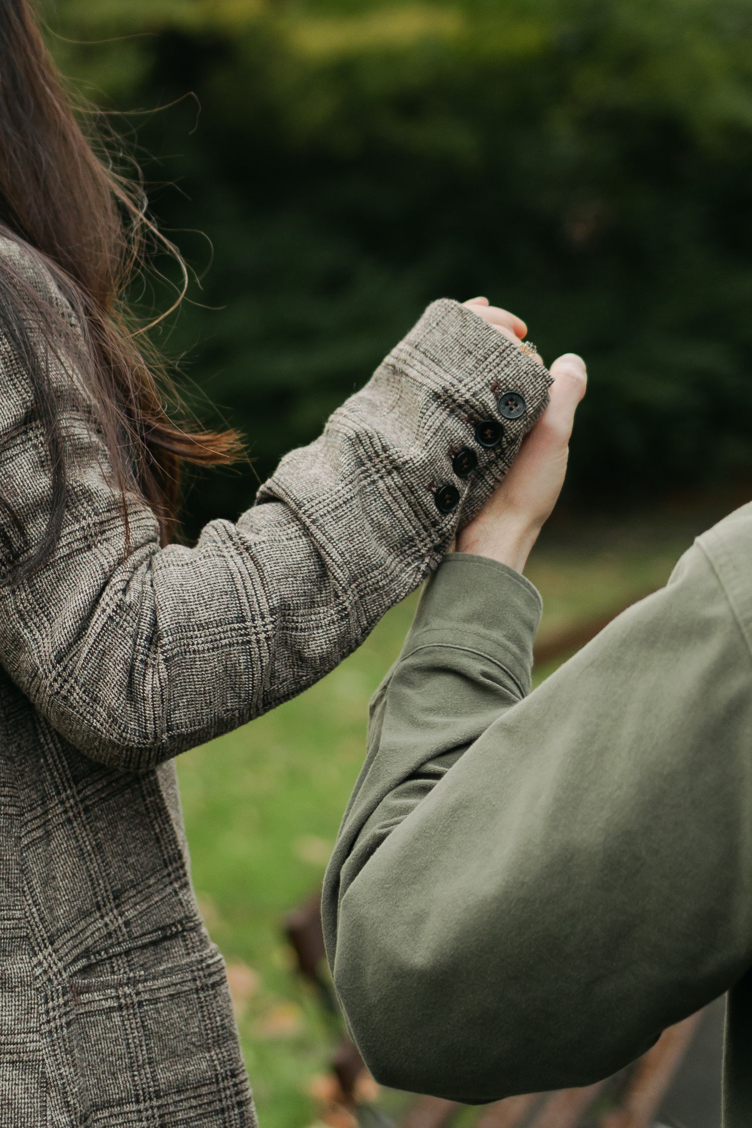 Two people holding hands outdoors, one wearing a grey plaid blazer and the other a sage green sweatshirt.