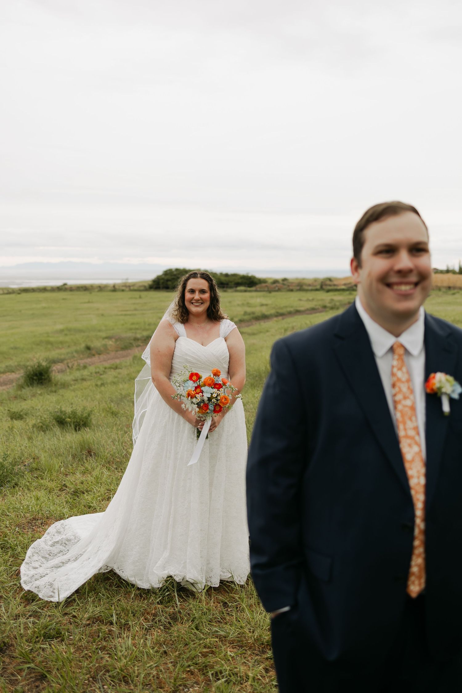 Bride in white gown holding orange bouquet stands in green field as groom in navy suit smiles in foreground.