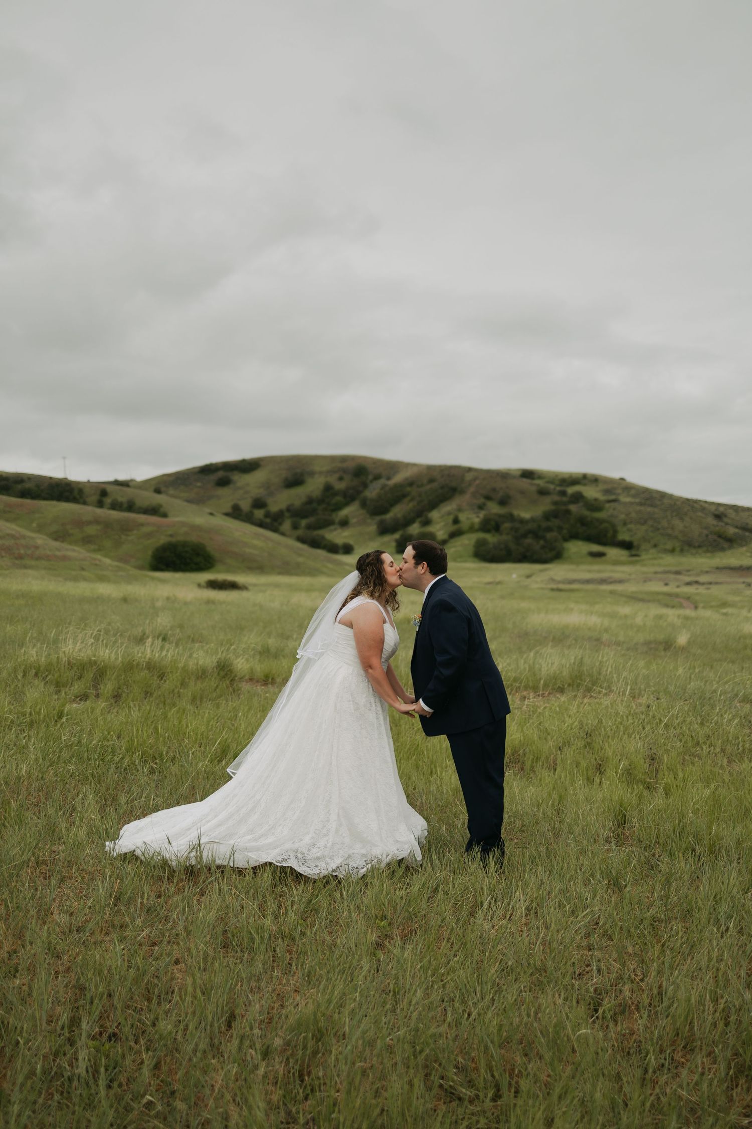 Bride and groom kissing in a lush green field with rolling hills, bride in white lace gown with veil.