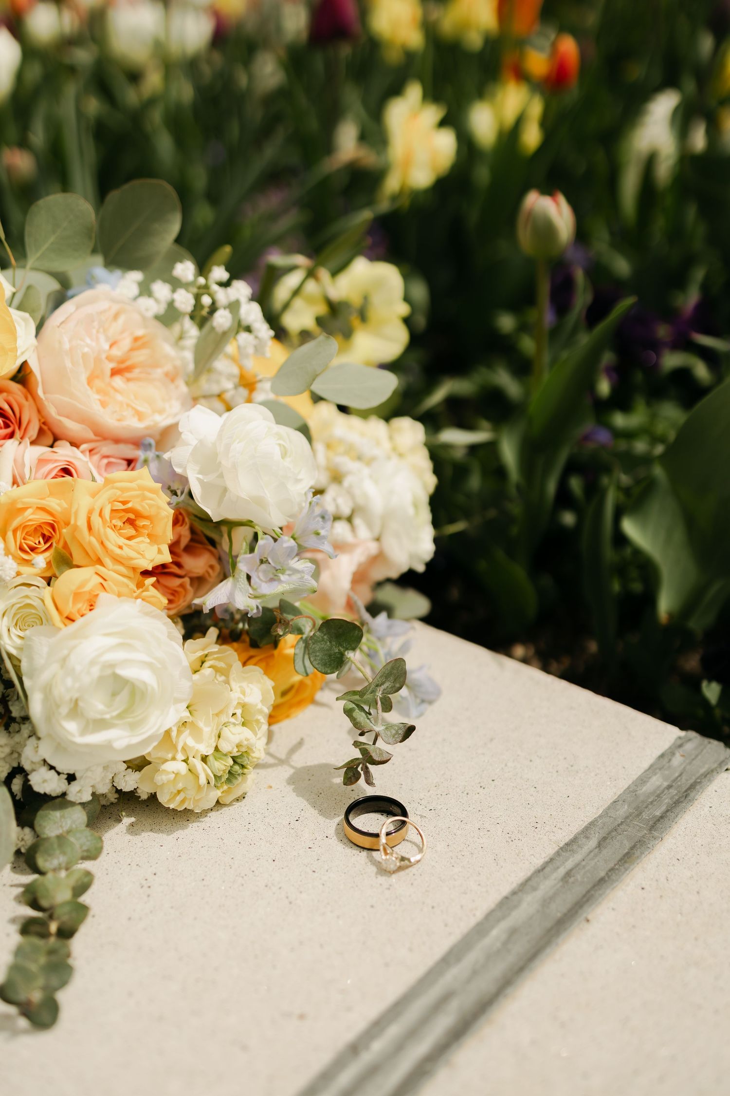 Wedding rings nestled among peach and white roses with delicate greenery on a white surface.