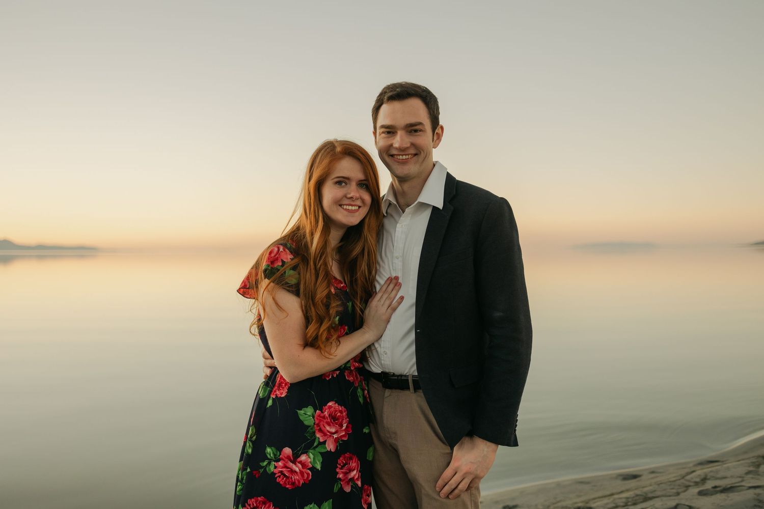 Romantic couple poses against sunset ocean backdrop in formal attire.