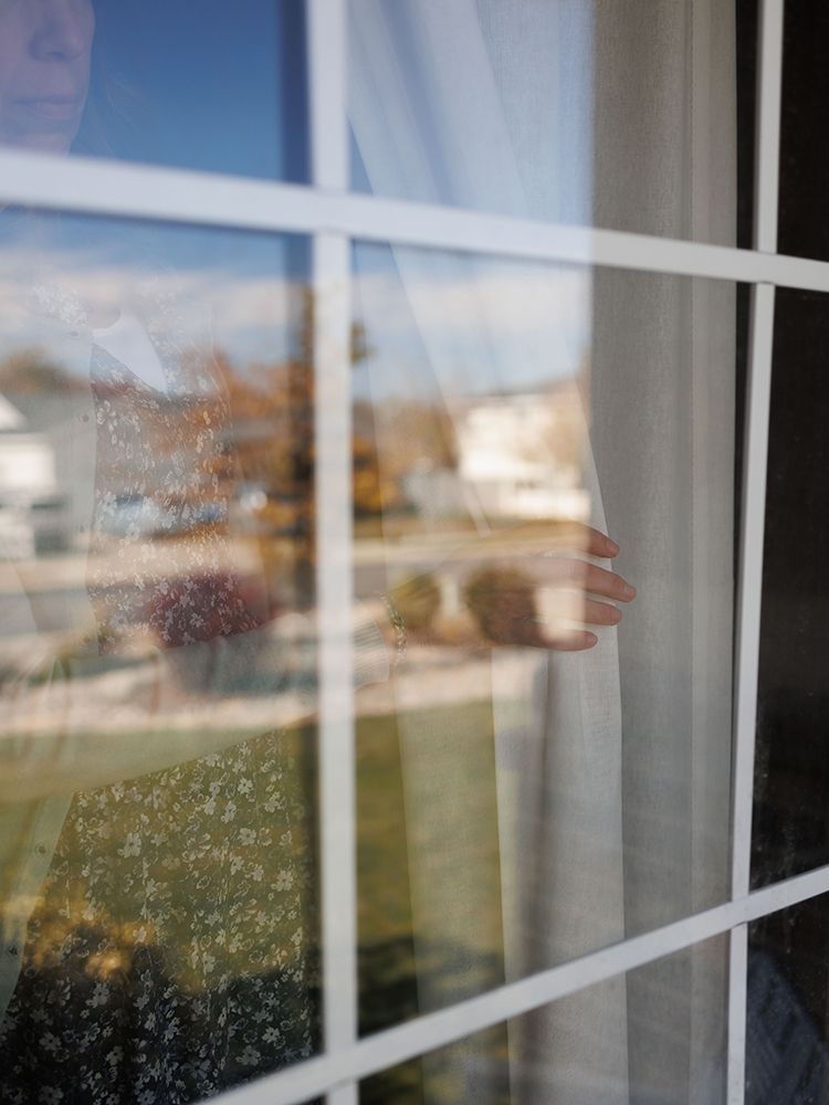 Person peering through a white-framed window with sheer curtains, reflecting a suburban neighborhood on a sunny day.