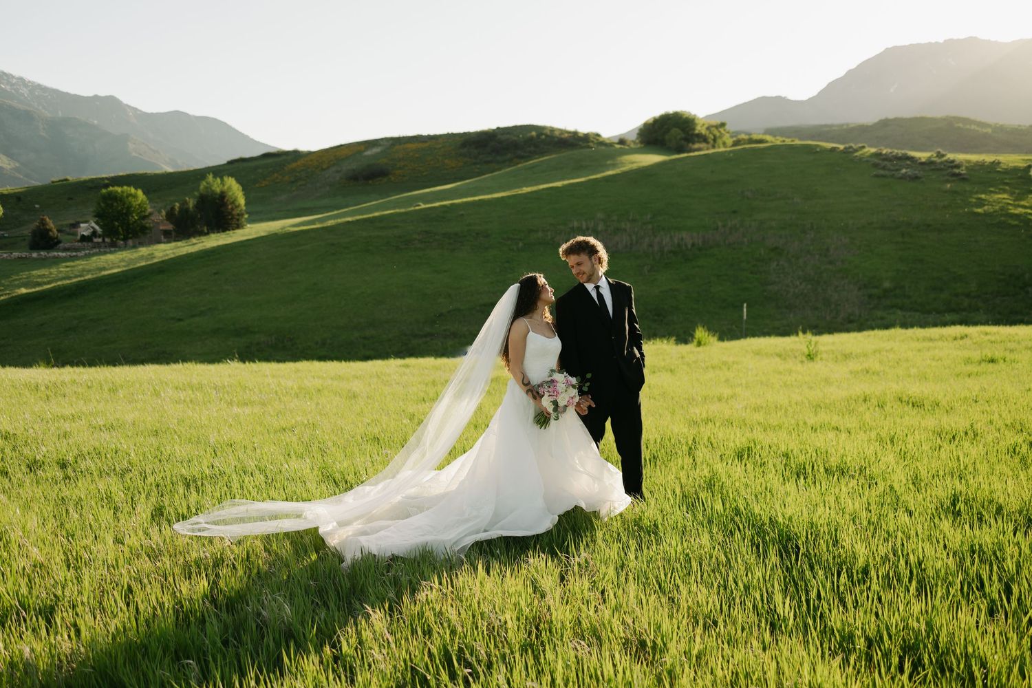Bride and groom walk hand in hand through rolling green hills at sunset.