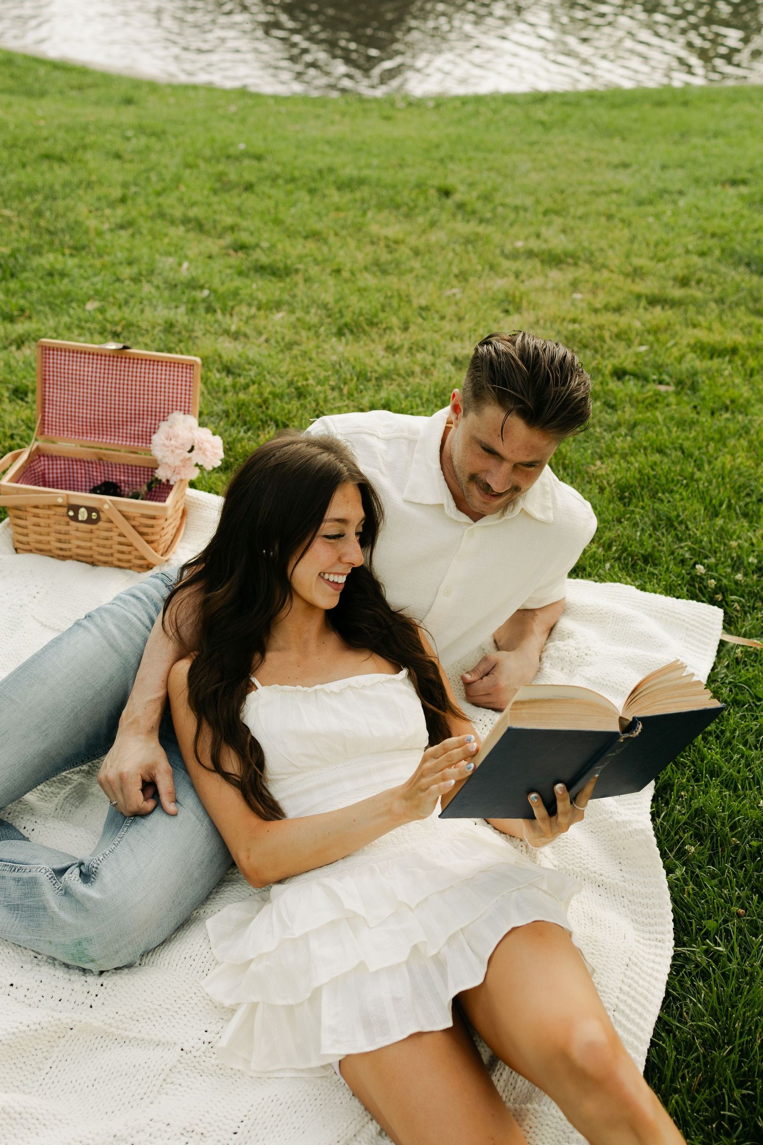 A couple enjoys reading books together during a summer picnic by the water, with a wicker basket nearby on a blanket.