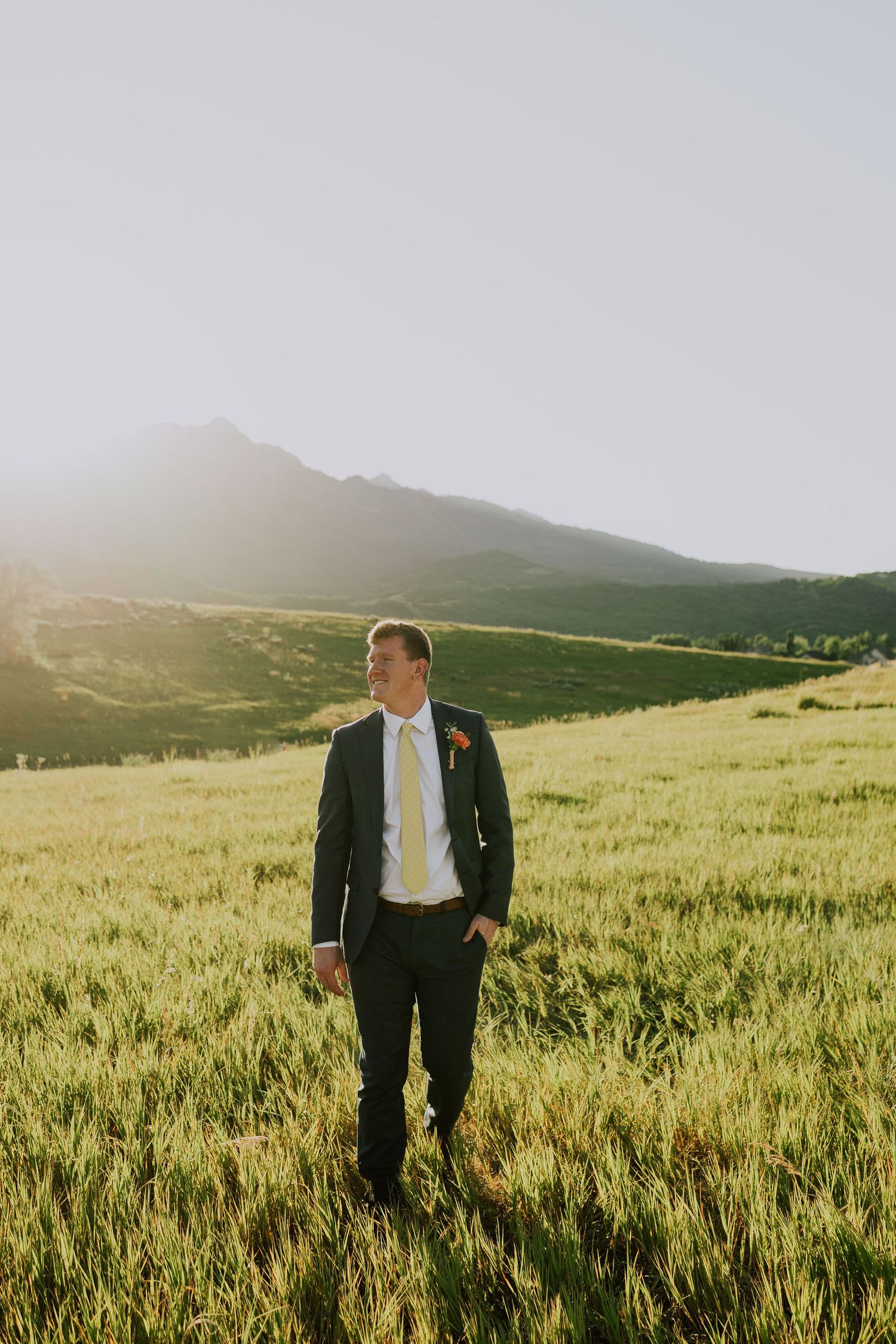 A person in formal attire stands in a golden-lit grassy meadow with mountains in the background.