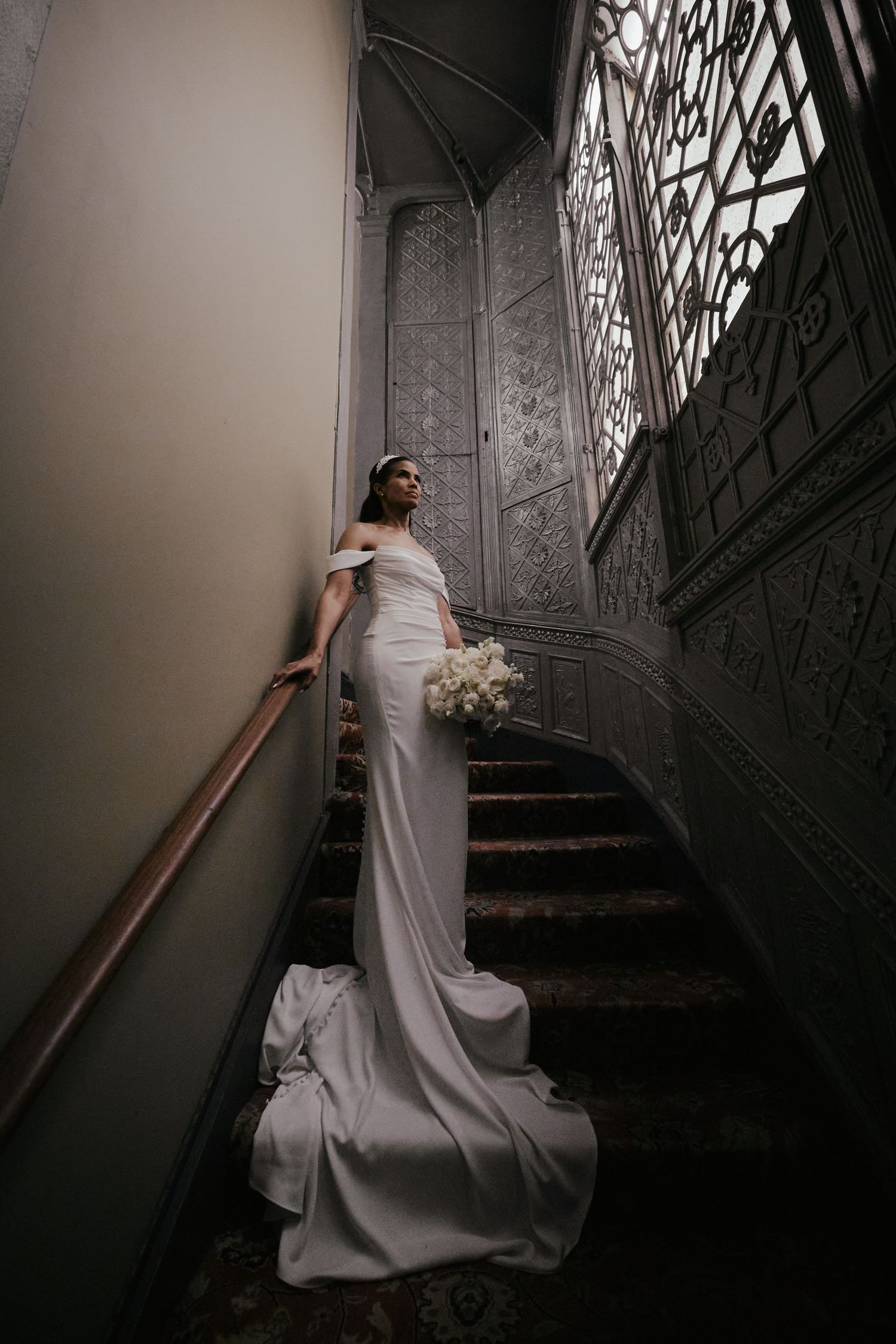 Bride in elegant white off-shoulder gown with train poses on ornate staircase holding bouquet.