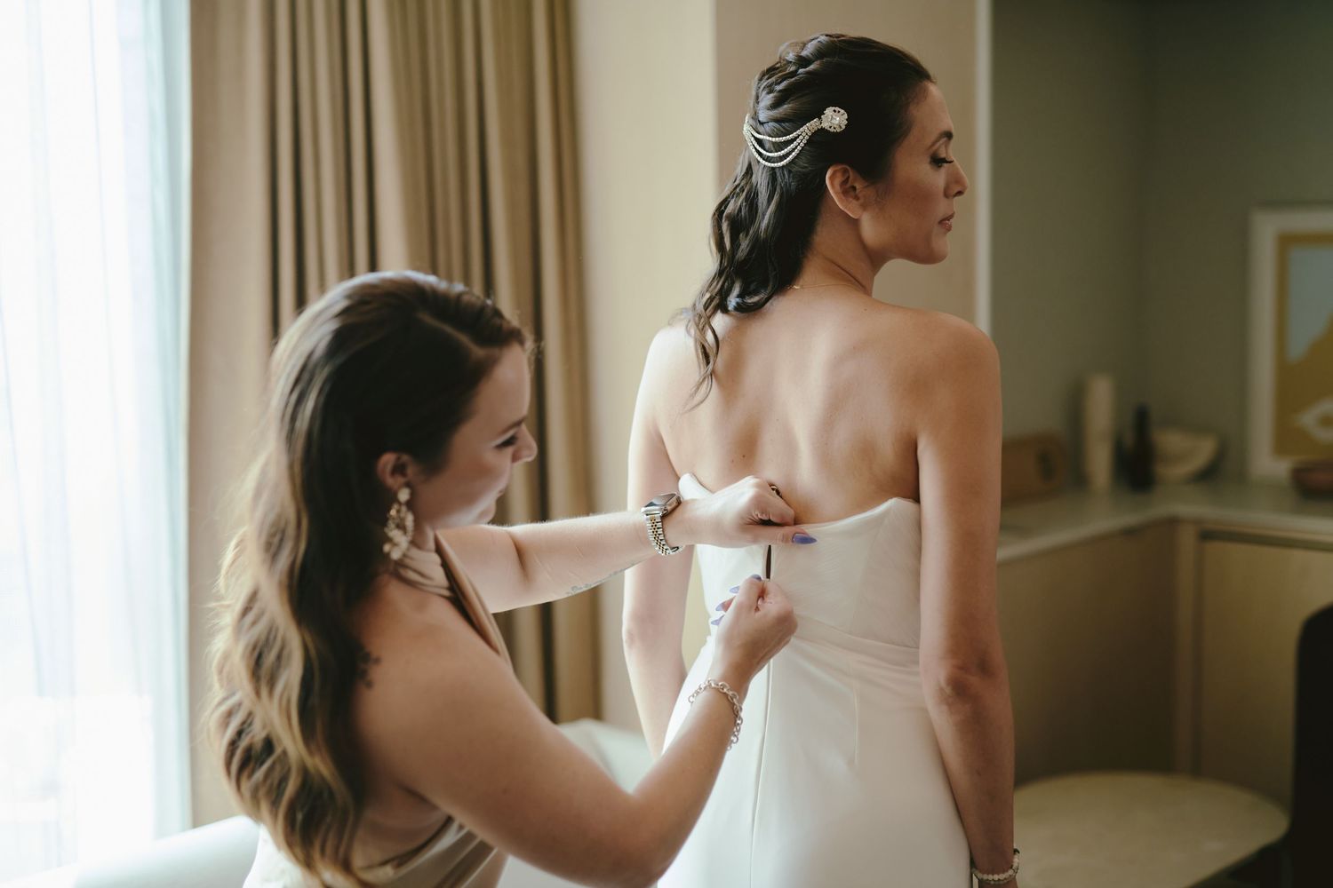 A bridesmaid helps zip up a white strapless wedding gown in a sunlit room.