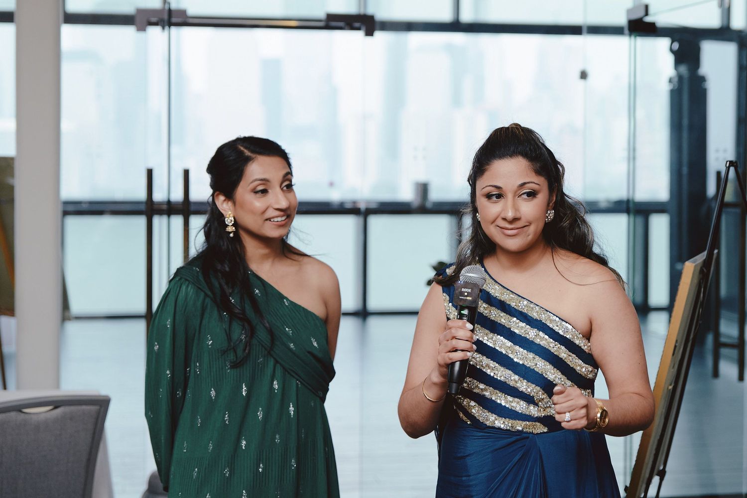 Two women in elegant evening gowns, one in dark green and one in blue with sparkles, smiling at an indoor event.