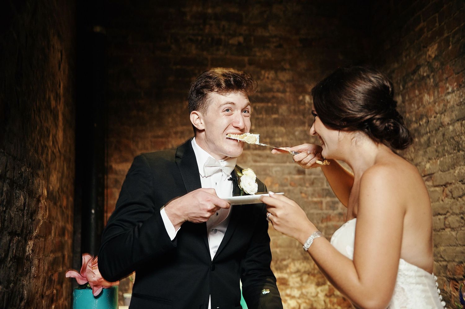 Newlywed couple playfully feed each other cake at rustic brick venue during wedding reception.