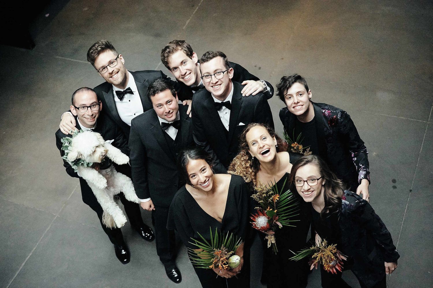 A wedding party group poses together in formal black attire on a dark floor while holding bouquets.