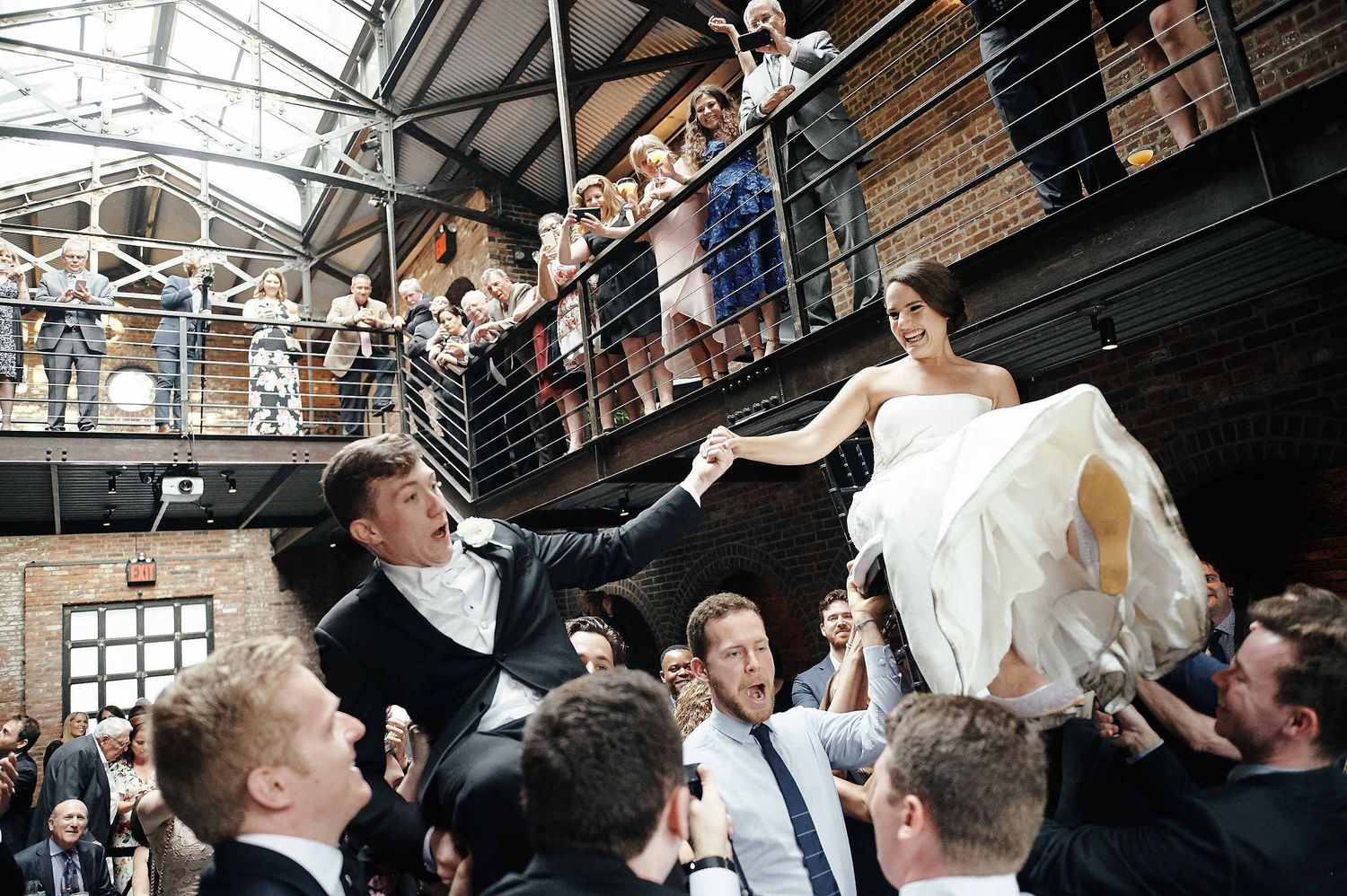 Wedding party celebrates from balcony as couple is hoisted up during joyful reception in industrial venue.