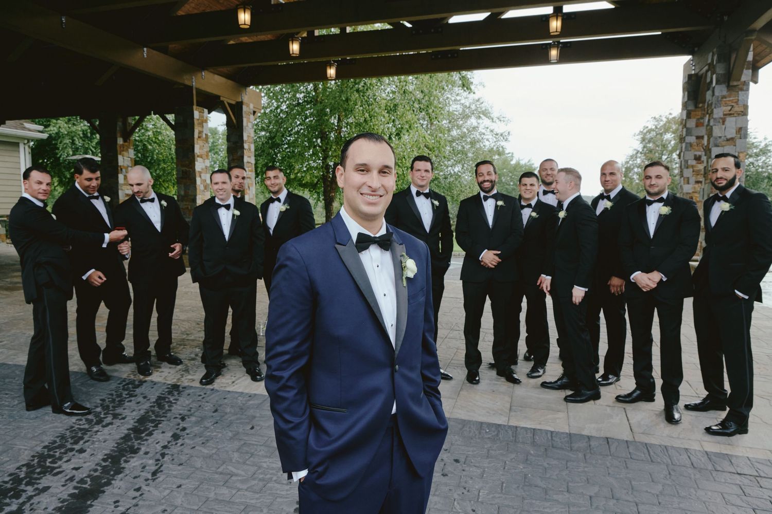 Groom in navy tuxedo stands in foreground while groomsmen in black suits gather behind at outdoor wedding venue.
