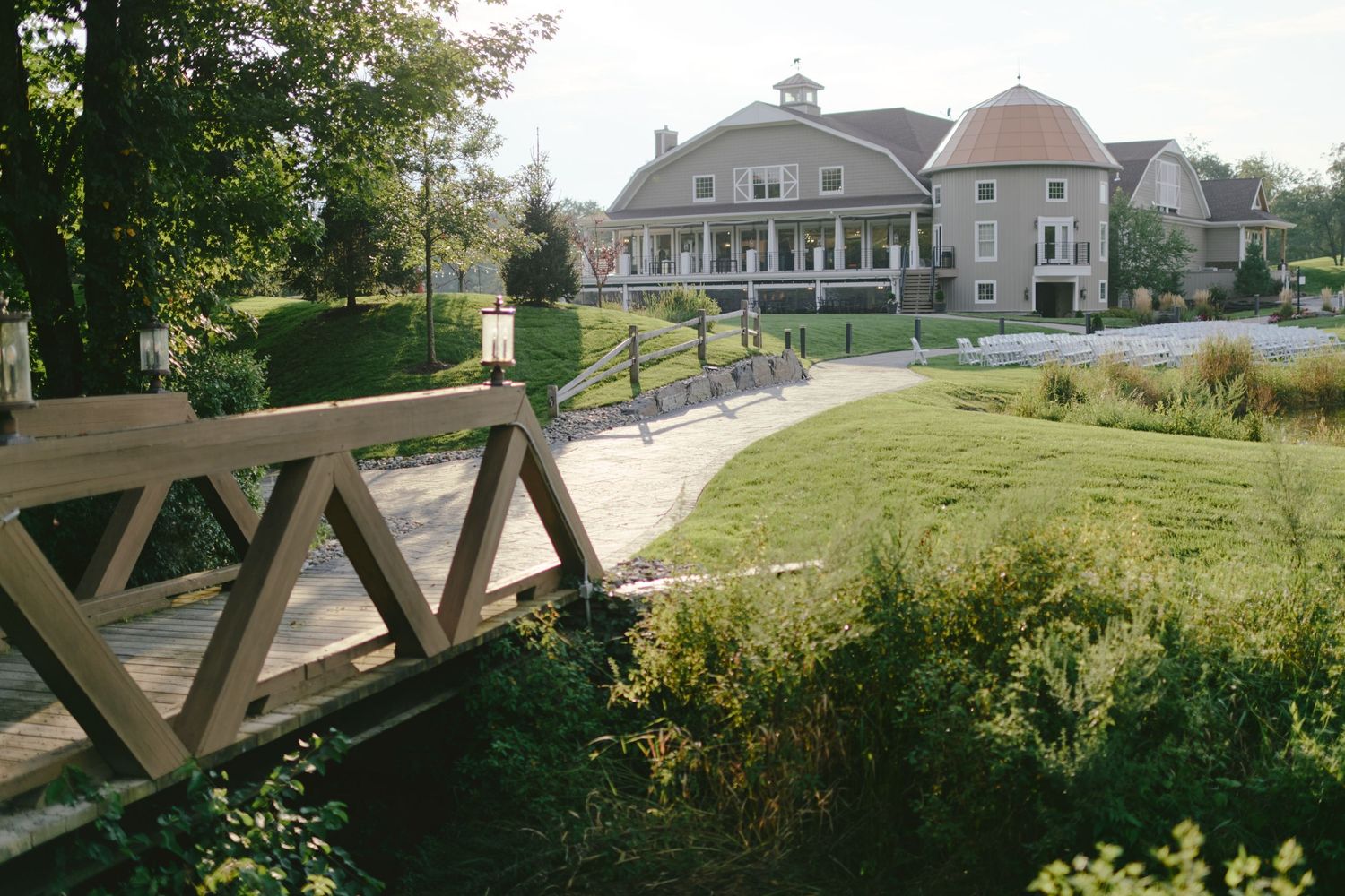 A wooden bridge overlooks a landscaped property with large gray houses and manicured lawns on a sunny day.