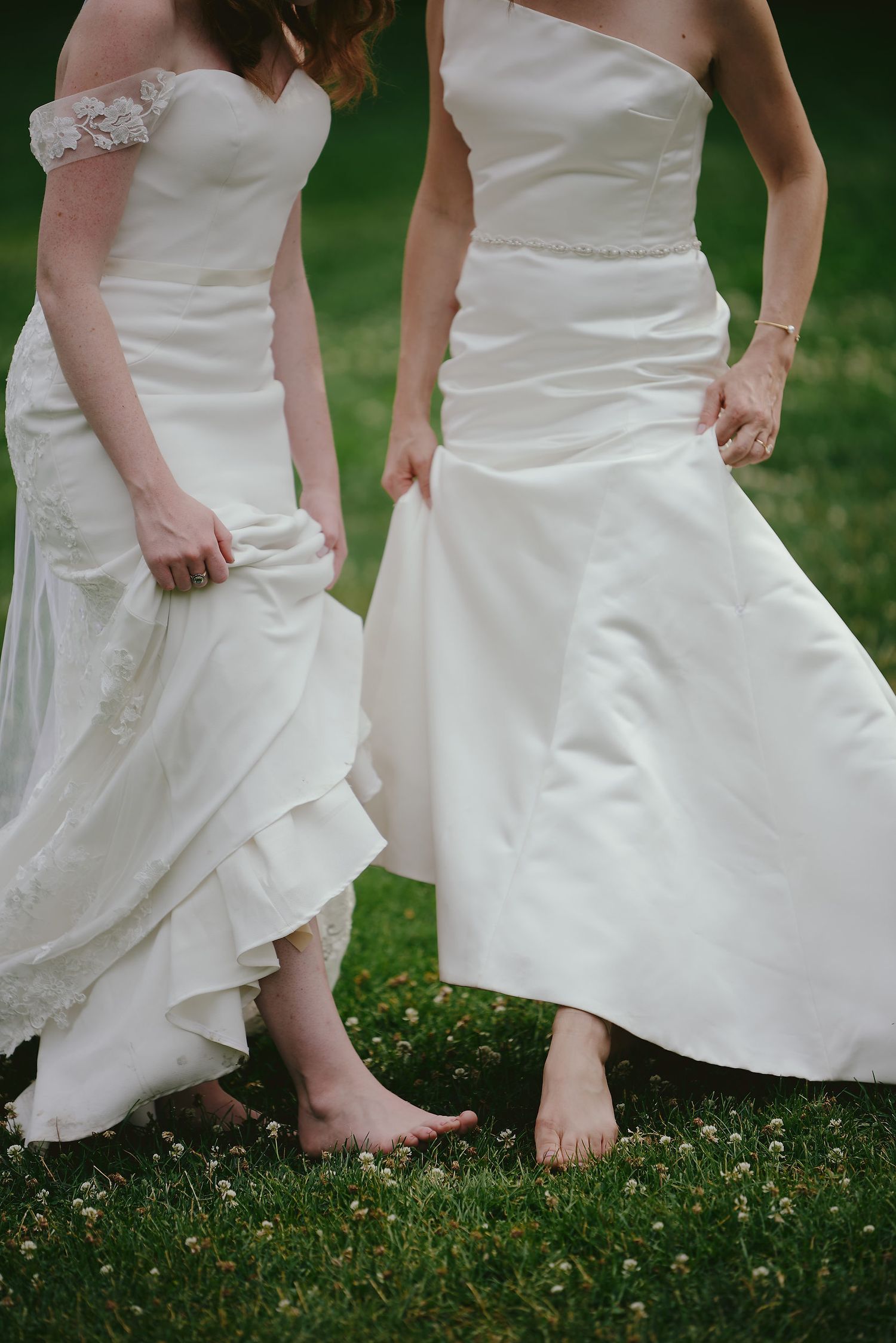 Two barefoot brides stand together on grass wearing white wedding dresses with delicate details and flowing skirts.