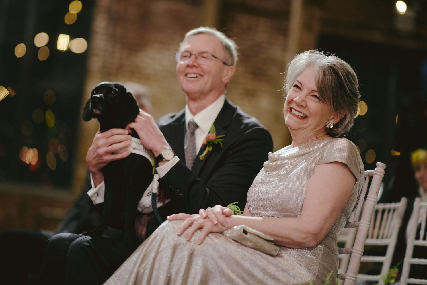 A couple sits together at a wedding ceremony while holding their black dog and laughing joyfully.