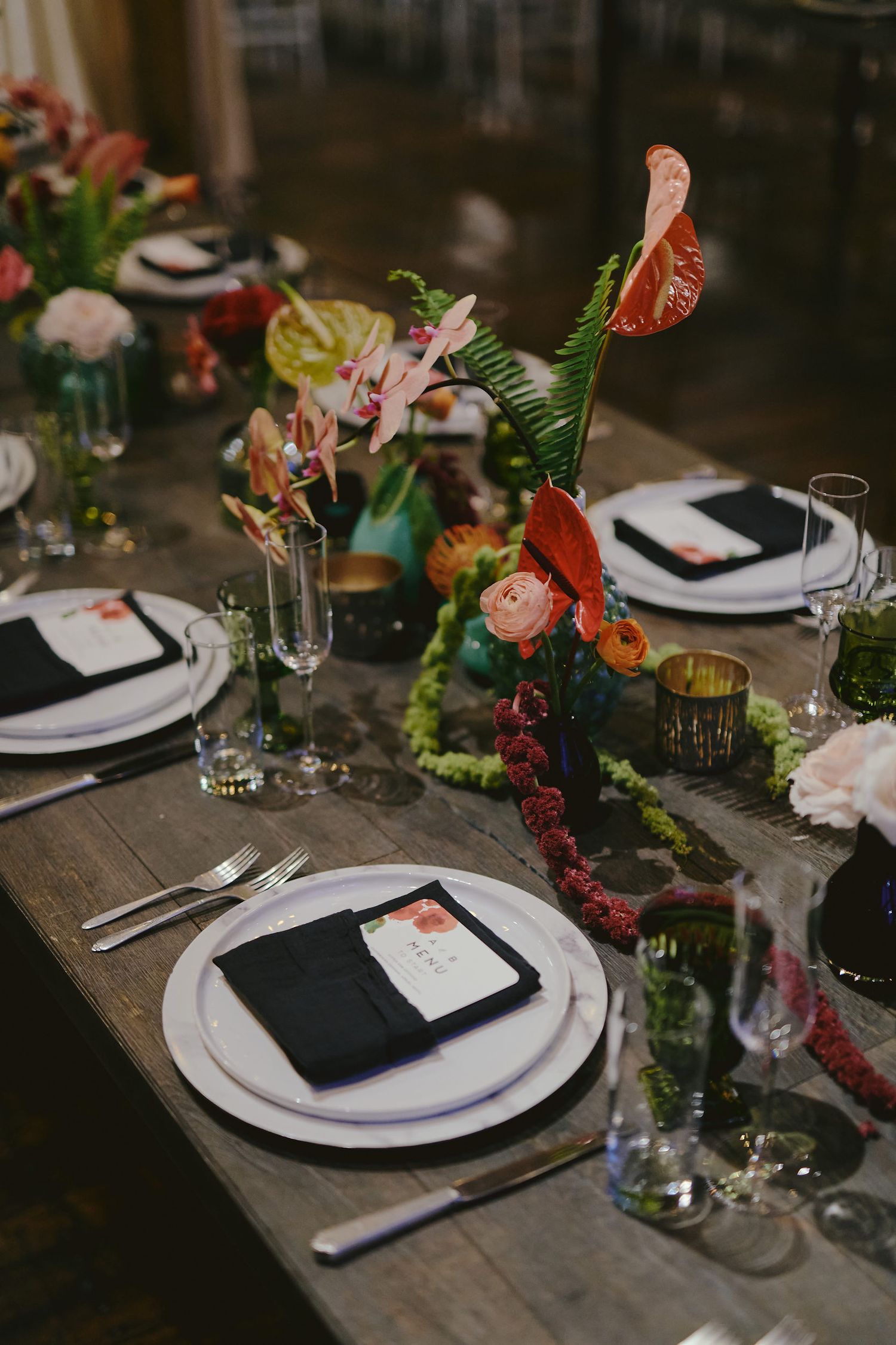 Elegant table setting with white plates, black napkins and colorful floral arrangements at a rustic wooden dining table.