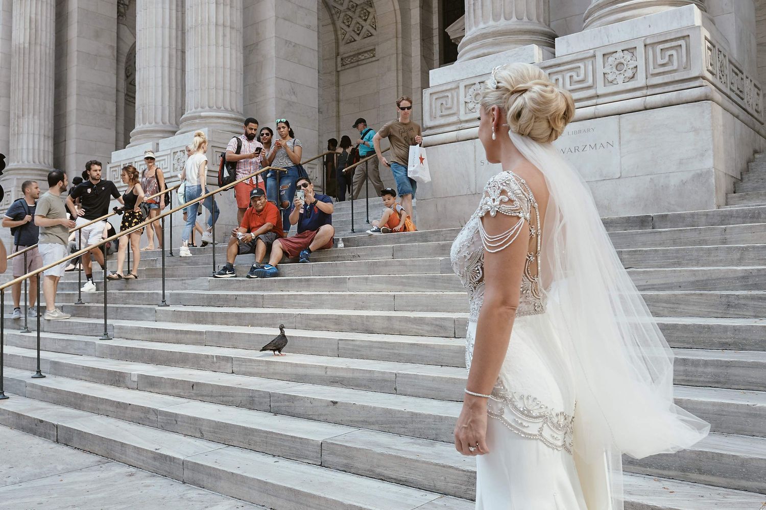 A bride in a lace gown and veil stands on grand marble steps while guests gather in the background near columns.