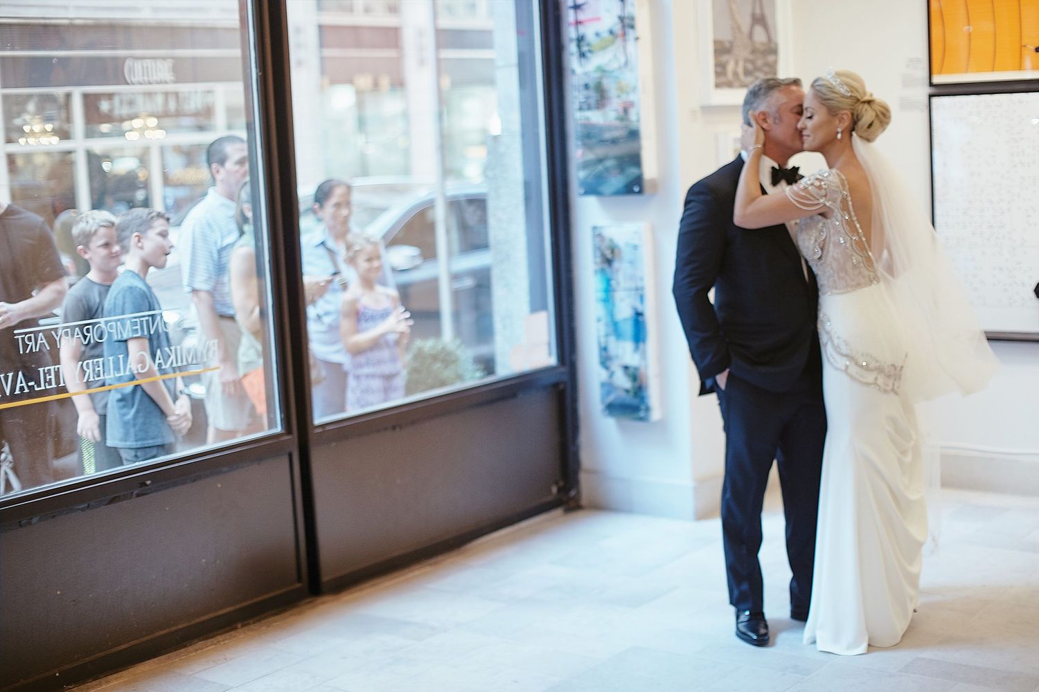A couple shares a romantic moment on their wedding day in front of large windows overlooking a busy street.