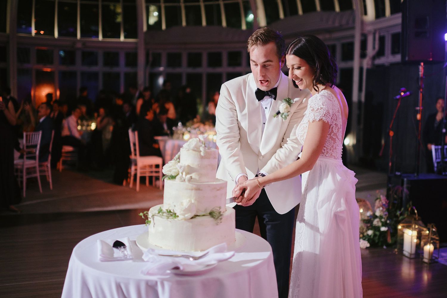 A couple in formal wedding attire cut their elegant white wedding cake together at an indoor reception venue.