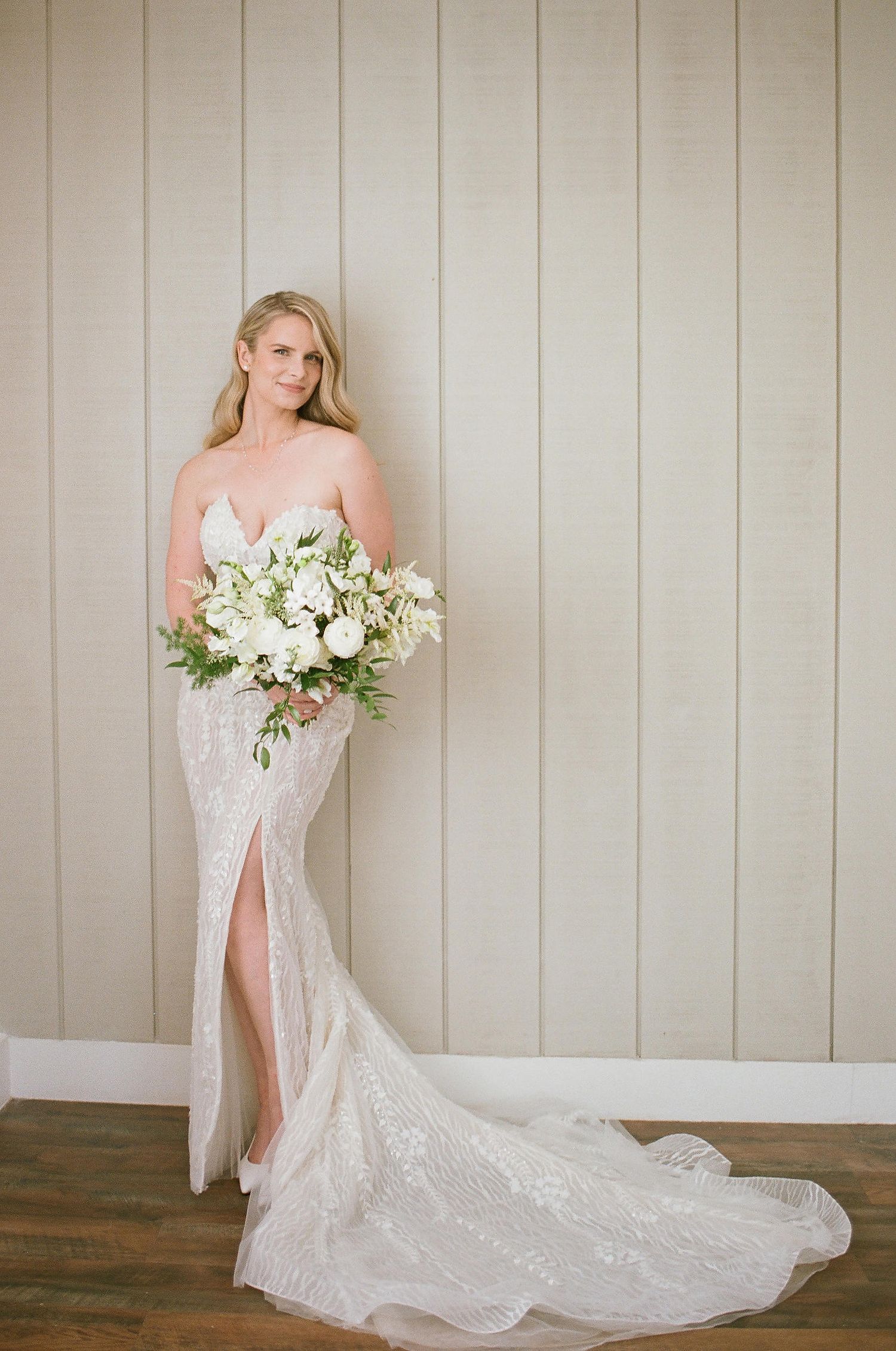 Elegant strapless lace wedding gown with side slit and long train, complemented by white and green floral bouquet.