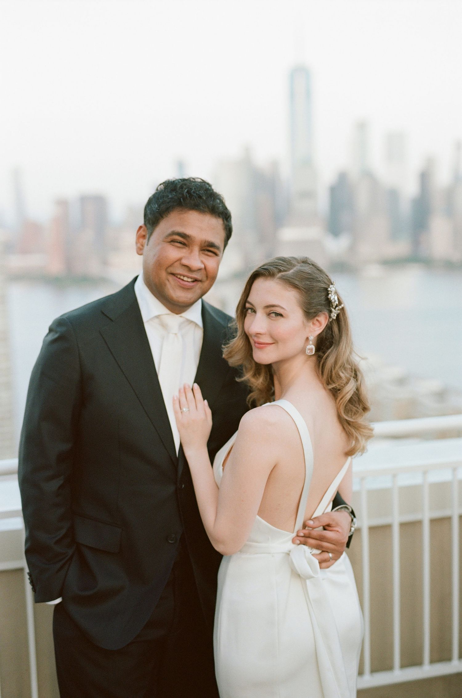 Wedding couple poses together on a terrace with a waterfront cityscape in the background.