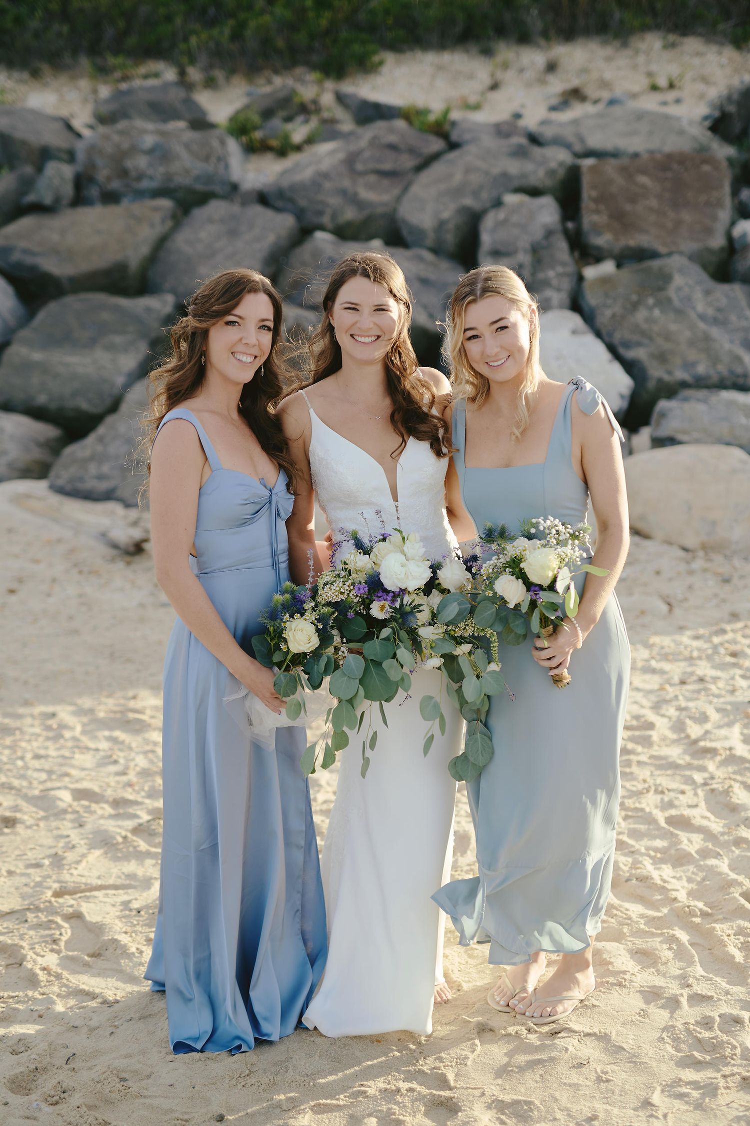 Three women in light blue bridesmaid dresses and white gown pose together on beach holding elegant bouquets.