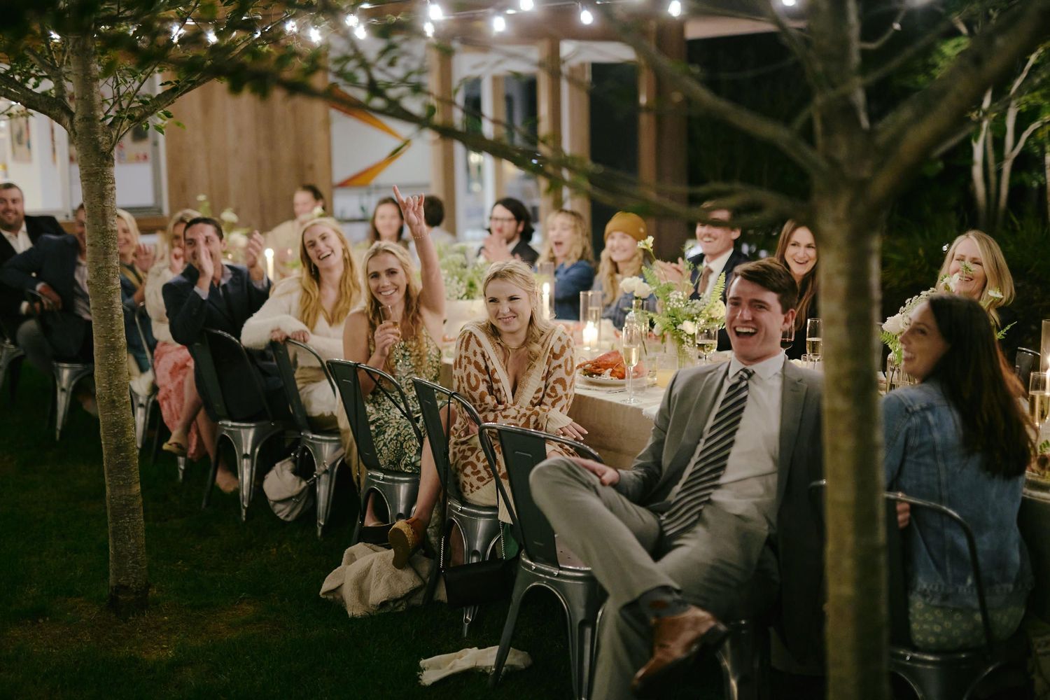 Wedding guests laugh and smile while seated at an outdoor evening reception under twinkling lights.