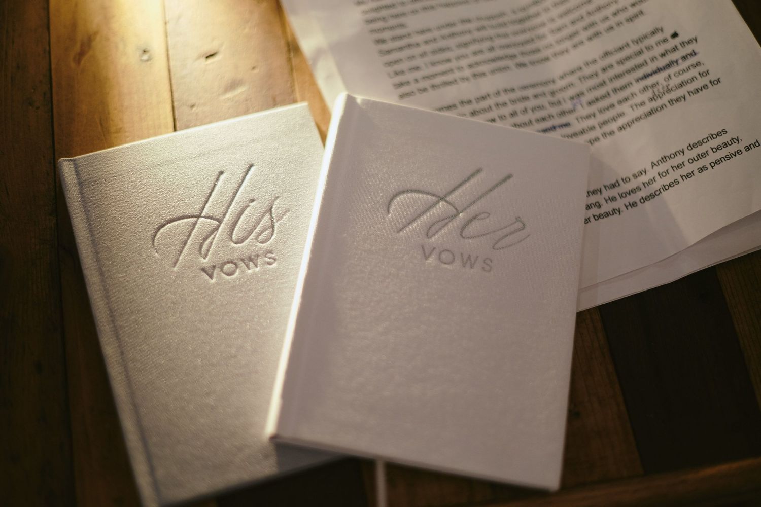 Two small white vows books labeled 'His' and 'Her' sit side by side on a wooden surface.