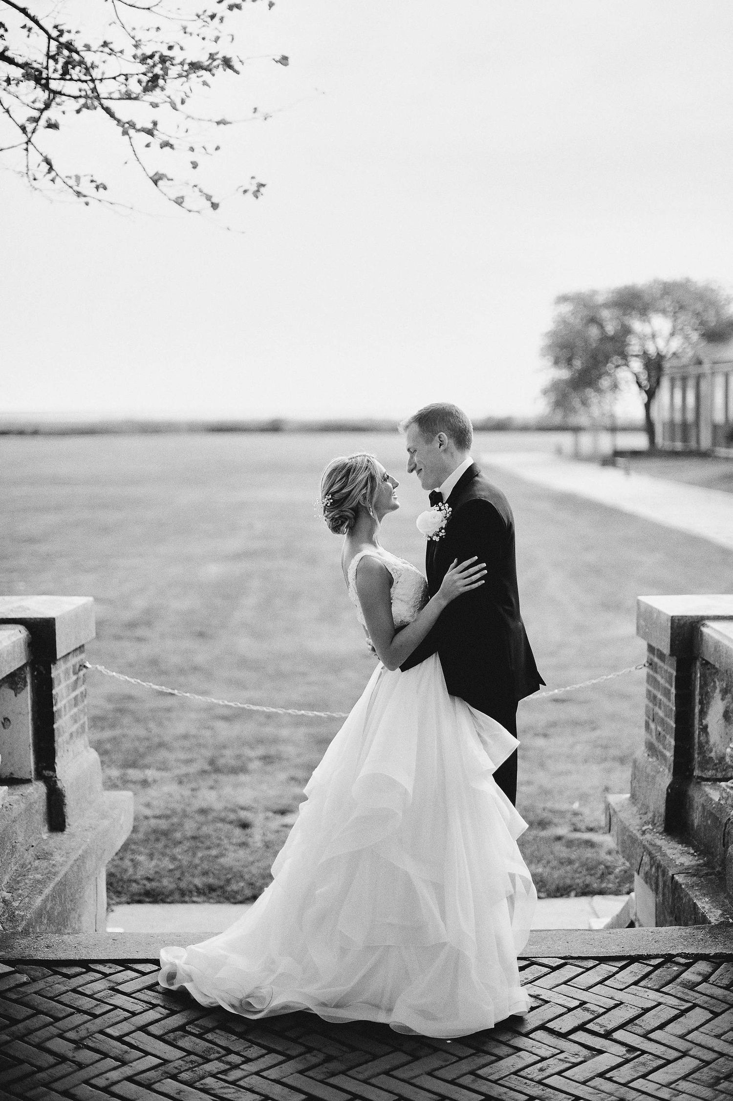A romantic black and white wedding photo of a couple embracing on a brick patio with open fields in the background.