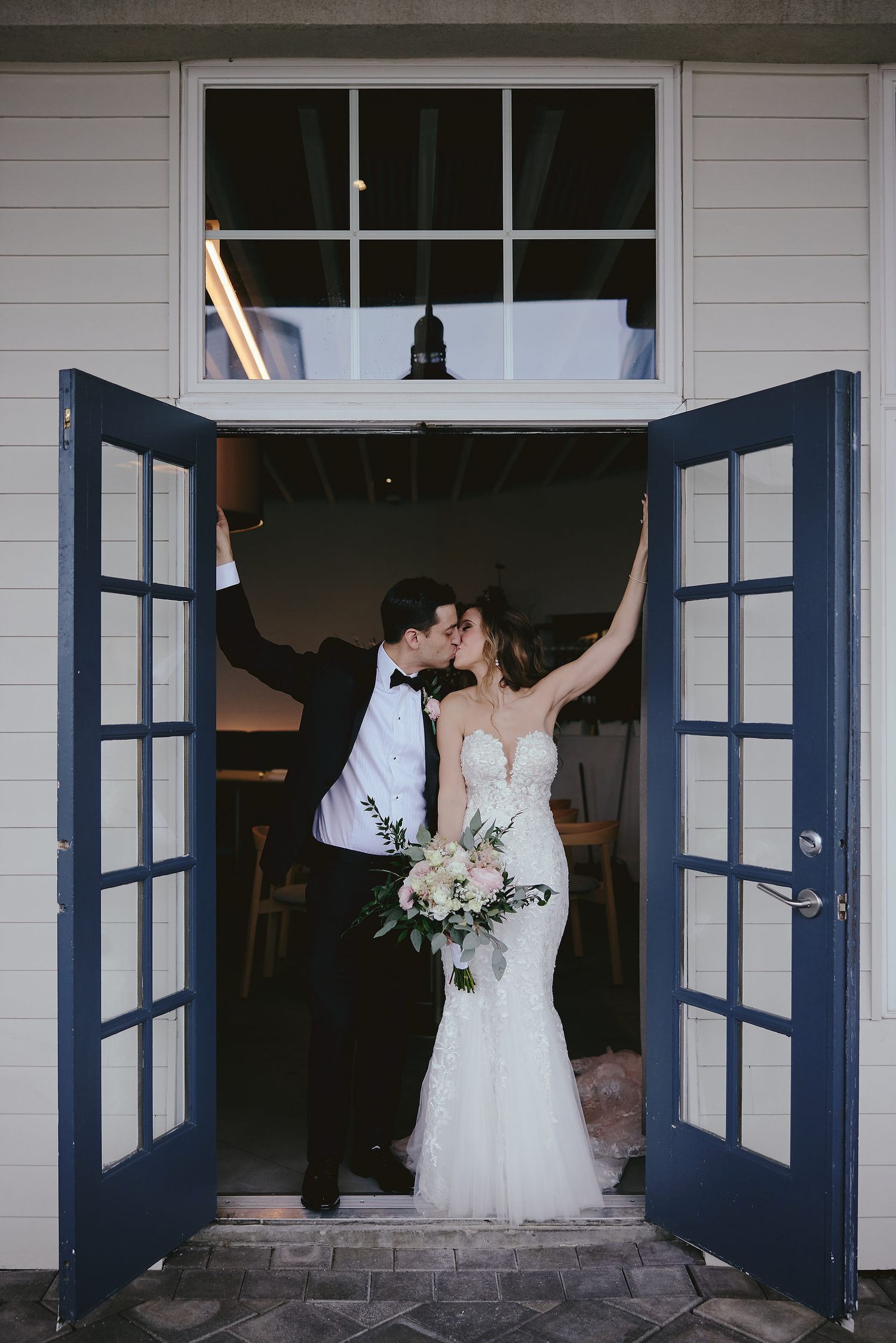 A romantic wedding moment captured in a doorway with navy blue French doors, featuring a white lace gown and formal attire.