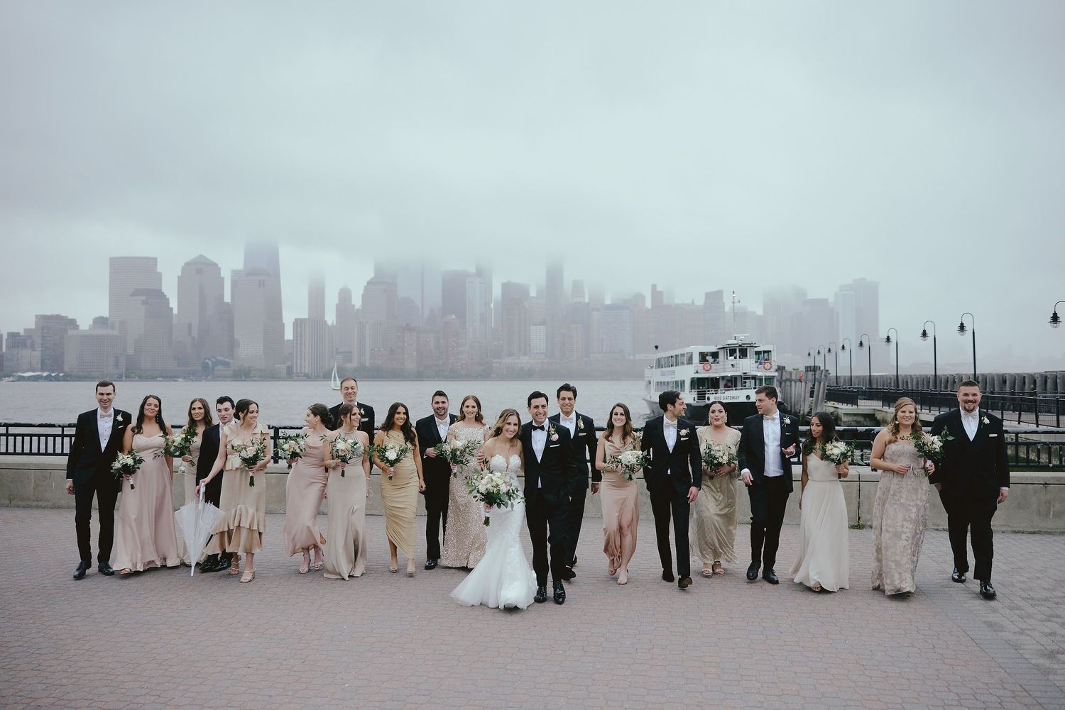 Wedding party poses together on waterfront promenade with foggy NYC skyline in background.