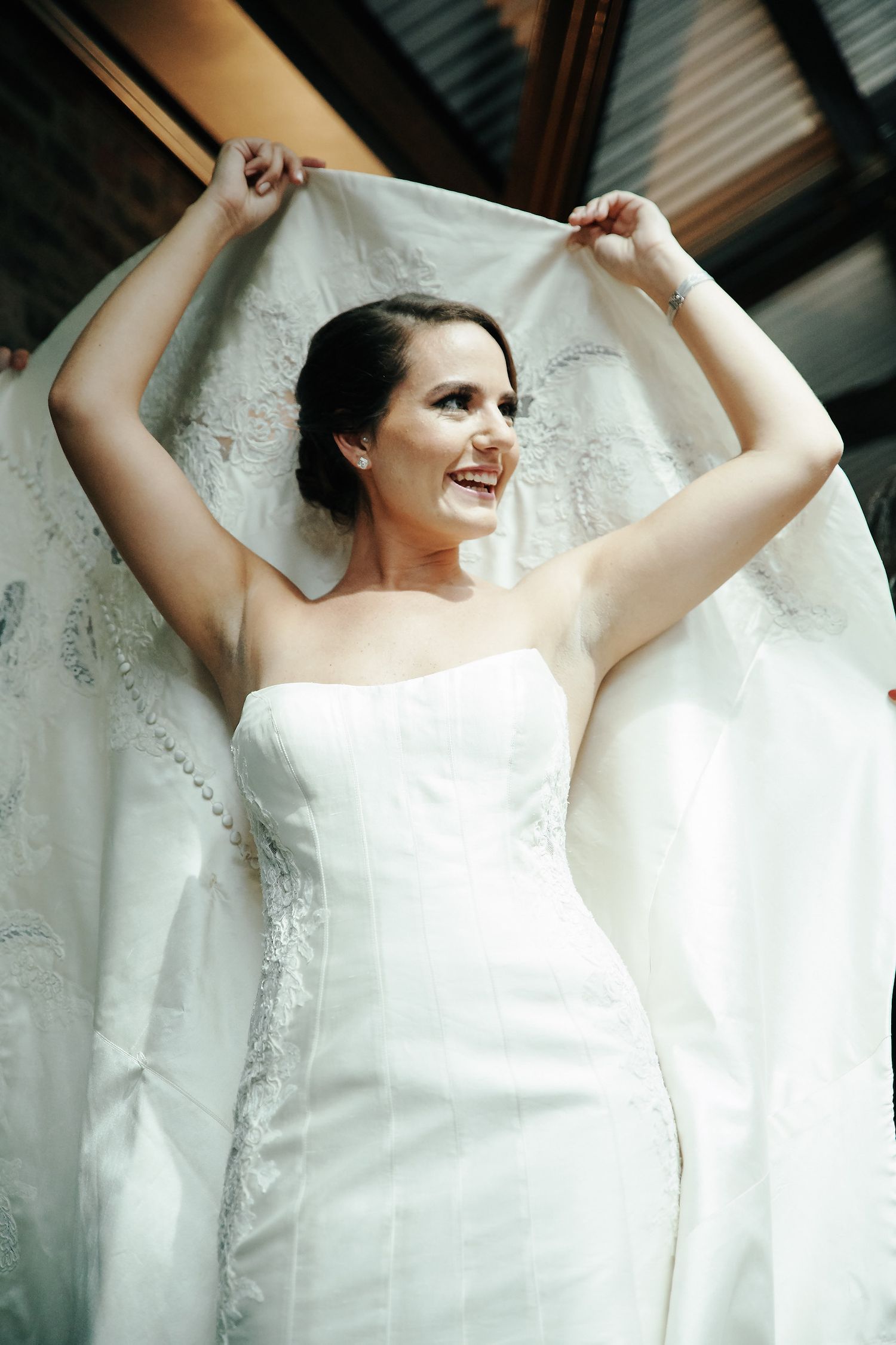 A joyful bride in a white strapless wedding dress holding up her veil in an elegant pose.