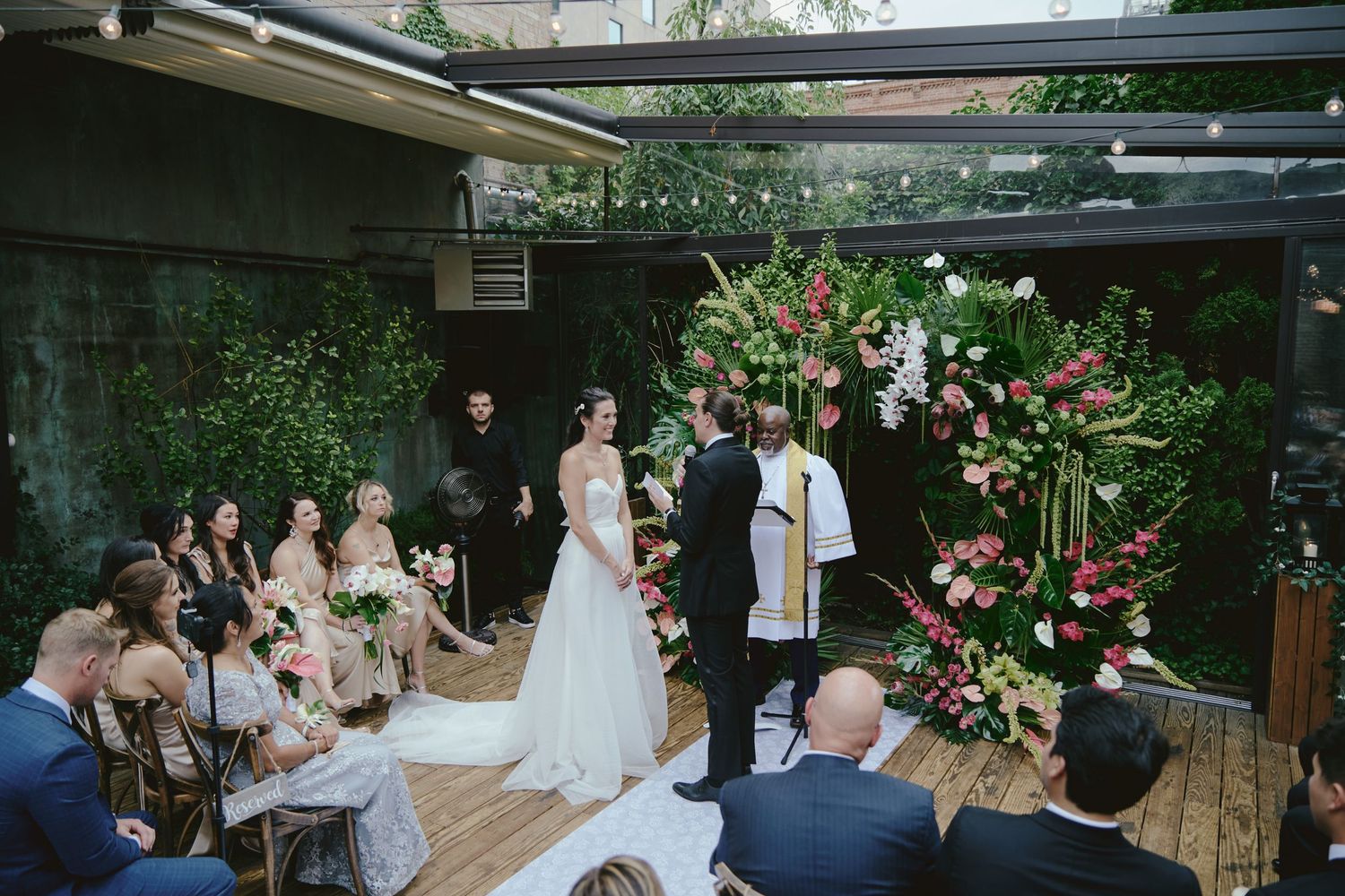 A small wedding ceremony taking place on a wooden deck under string lights with a large floral arrangement.