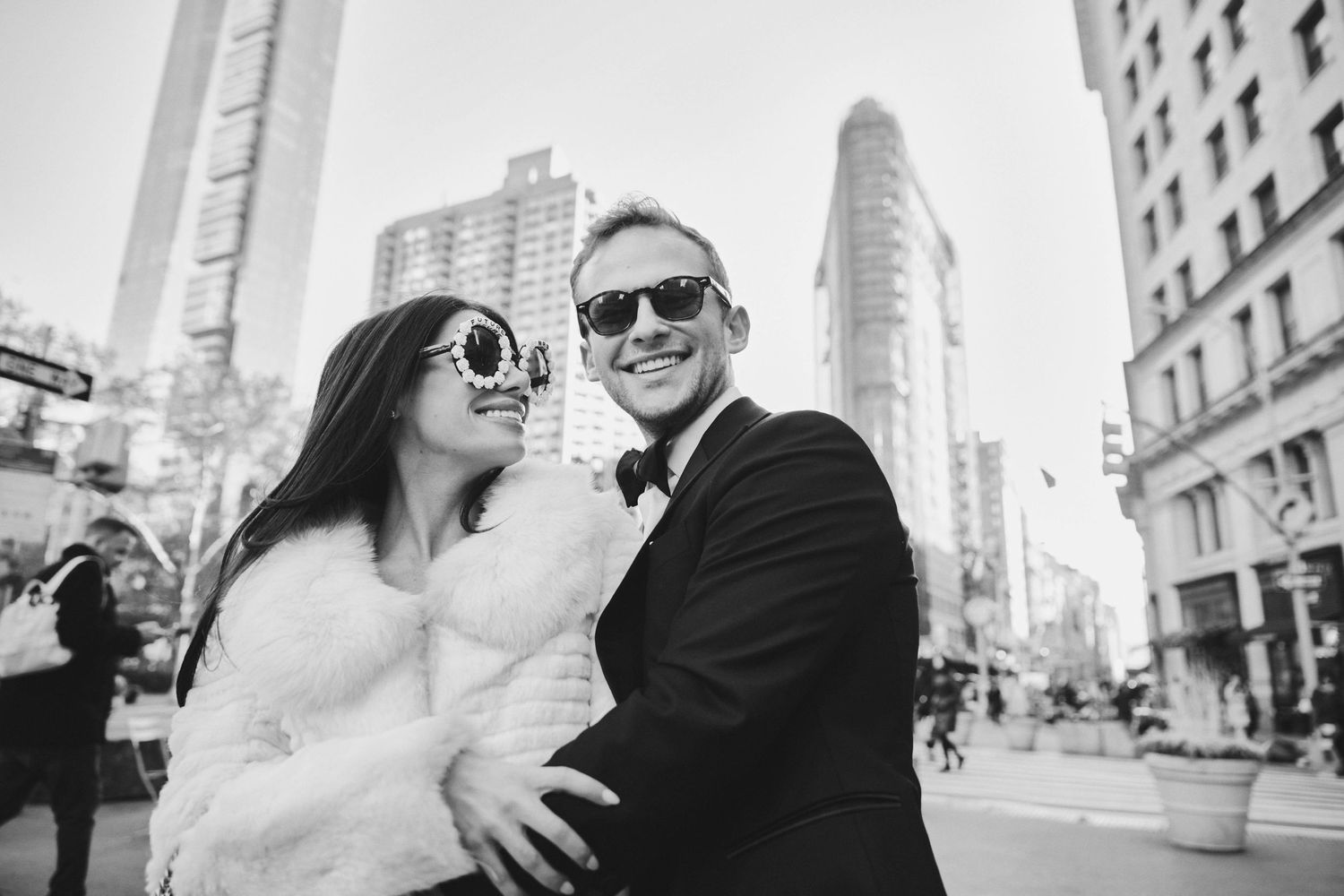 A couple in formal attire embraces while standing on a New York City street with tall buildings in the background.