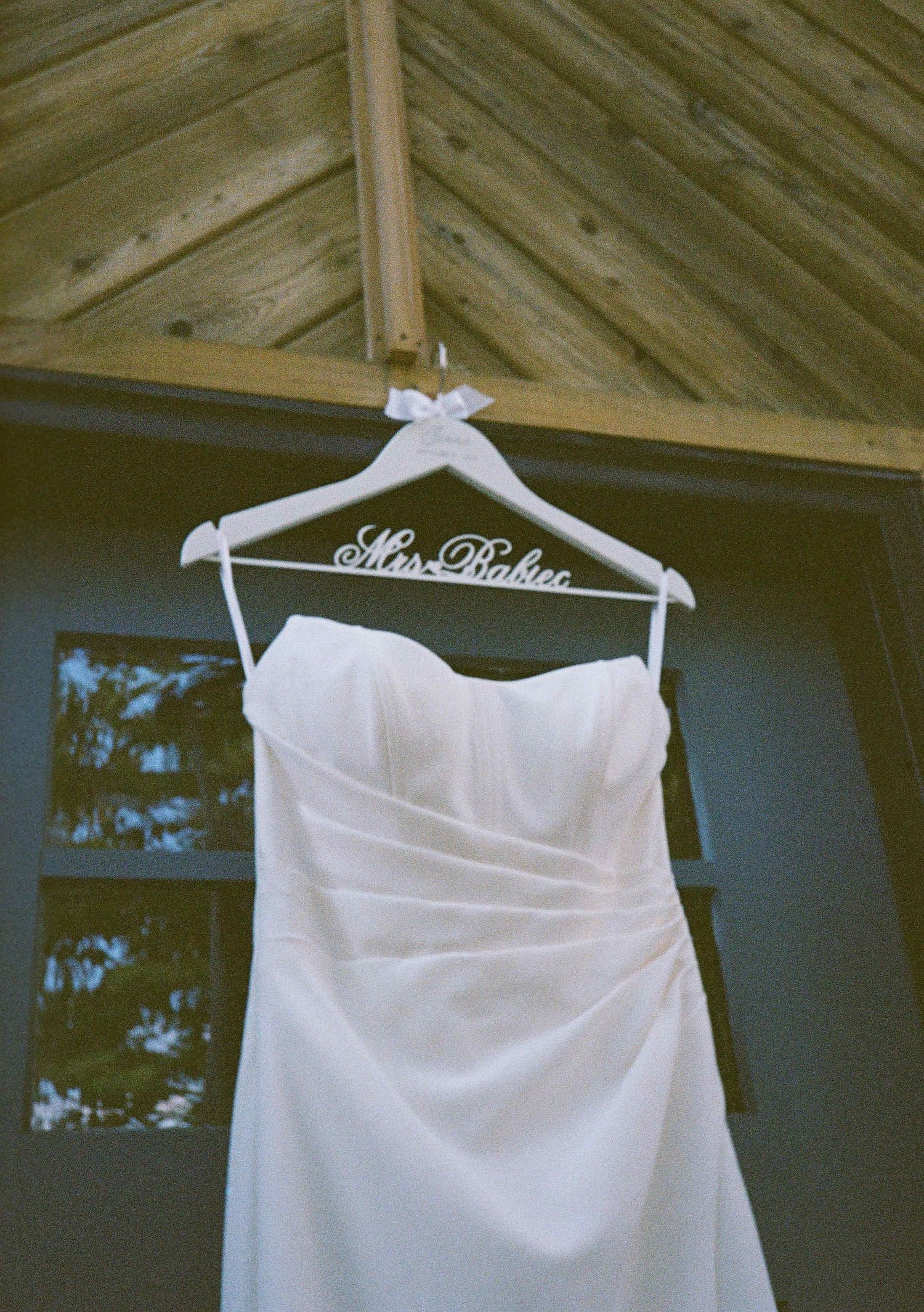 A white wedding dress hangs on a customized Mrs. Richter hanger against a wooden wall backdrop with natural lighting.