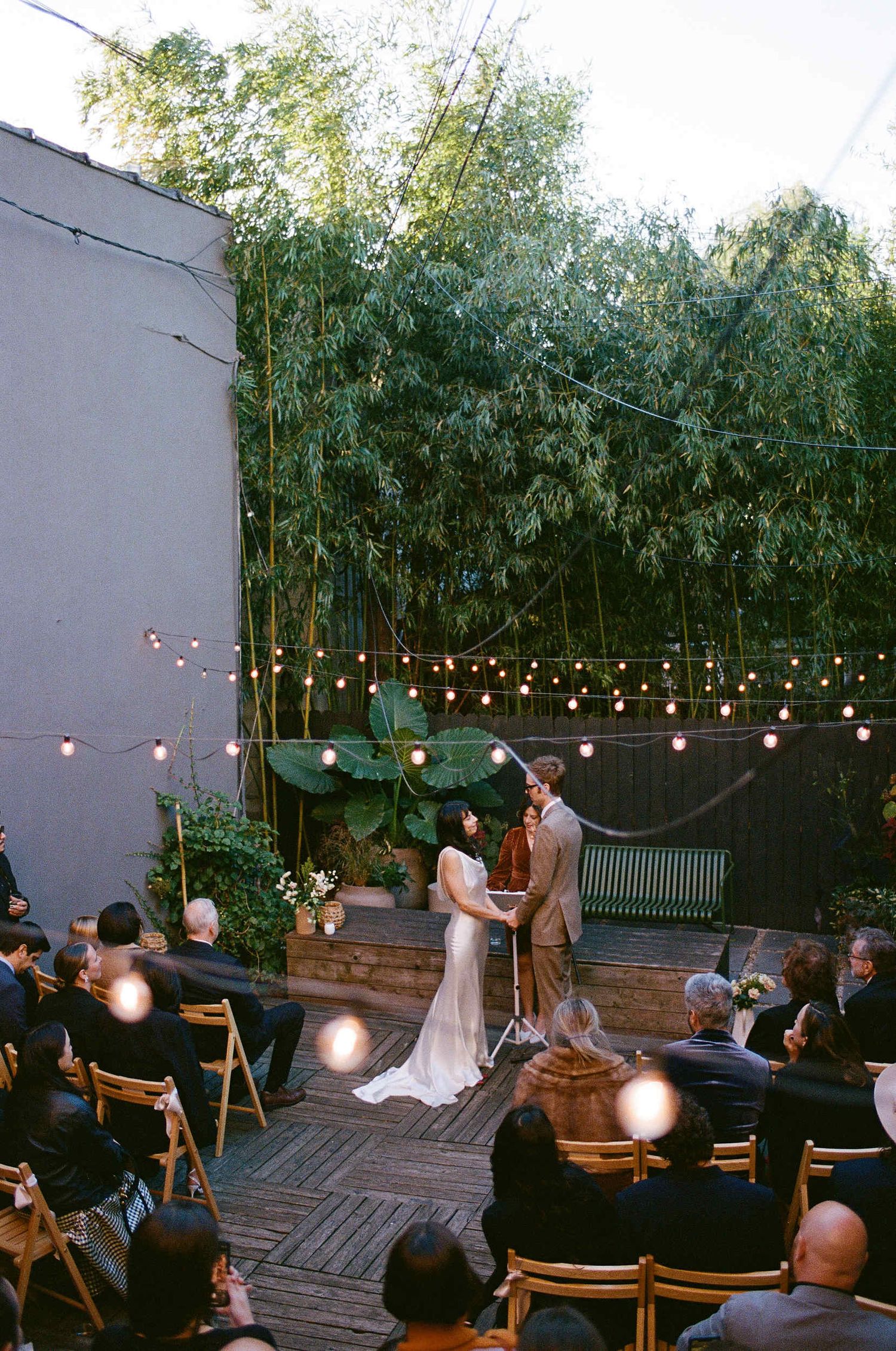 An intimate outdoor wedding ceremony under string lights with bamboo backdrop and guests seated on wooden chairs.