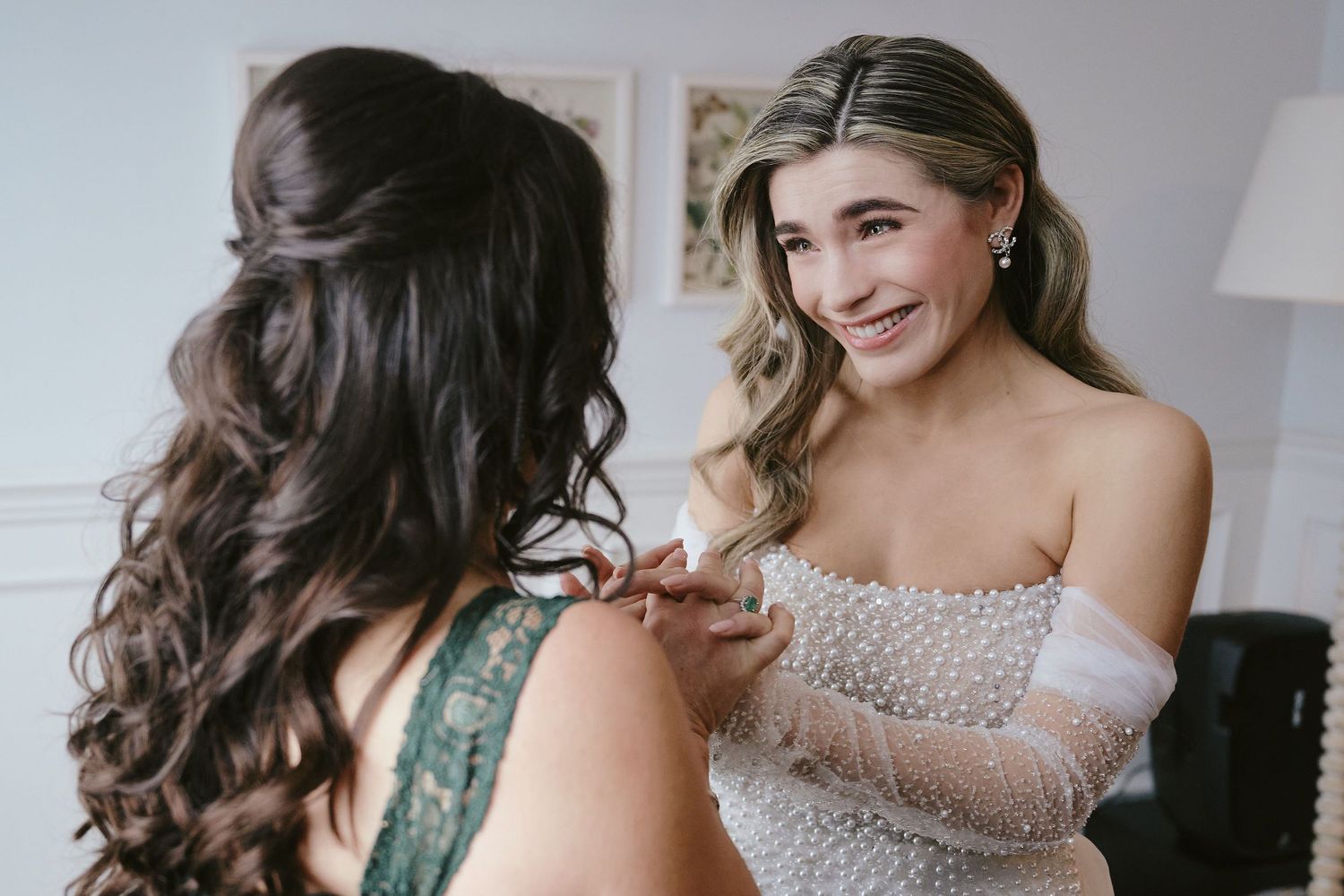 Two women share a joyful moment while getting ready, one wearing a strapless gown and the other in green lace.